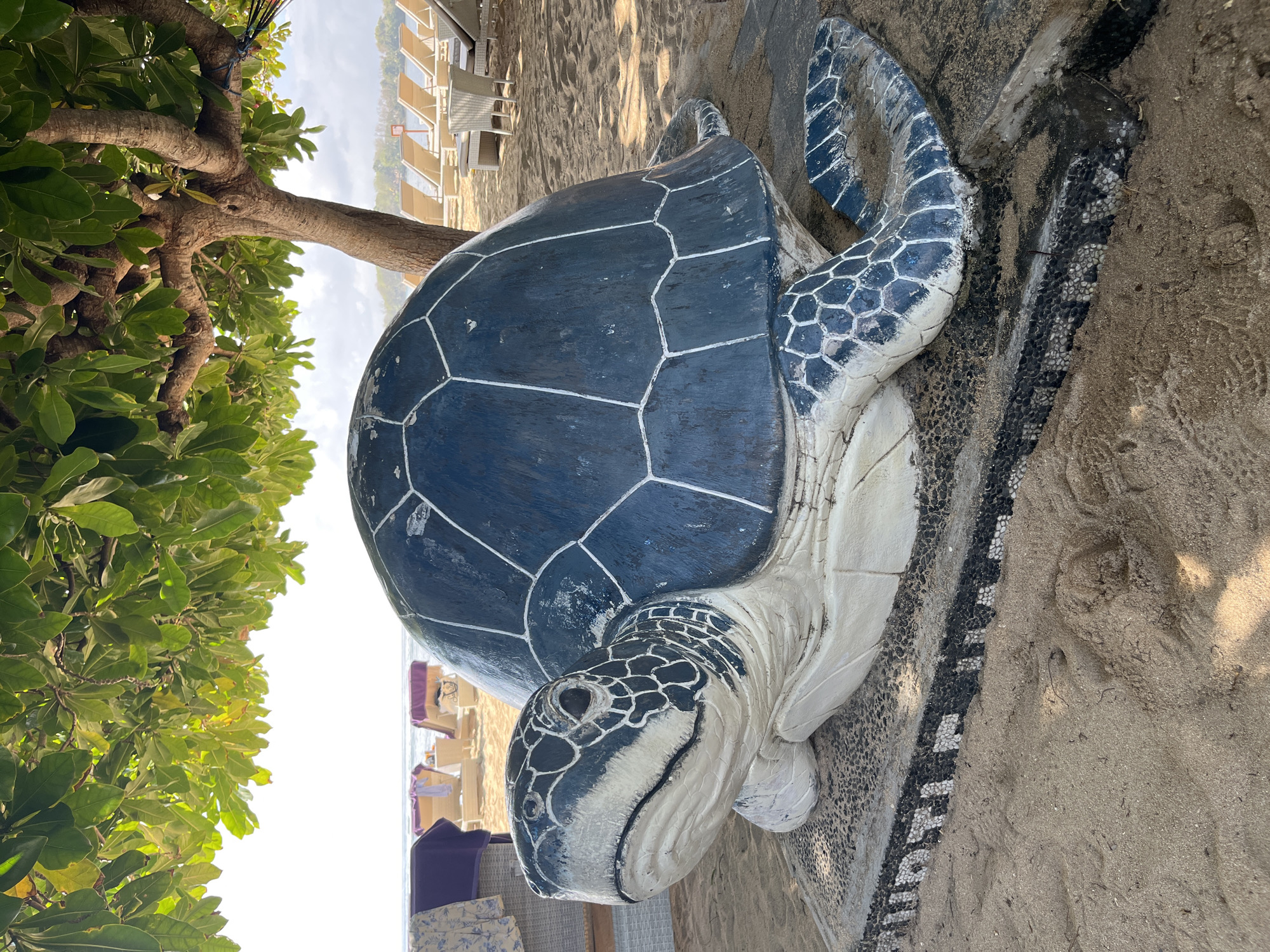 The image shows a large sculpture of a sea turtle on a sandy beach. The turtle is predominantly dark blue with white lines delineating its shell segments, and its head and flippers are white with blue patterned details. The sculpture is positioned on the sand, with some areas around it decorated with small black and white pebbles. To the left of the turtle, a tree with lush green leaves is visible, its branches extending downwards.
In the background, a resort or hotel with multiple floors and balconies can be seen, along with rows of empty lounge chairs and umbrellas on the beach. The sky is bright and appears to be clear, suggesting a sunny day. The overall scene depicts a tropical beach environment, likely a tourist resort. No people are visible in the image. The time of day appears to be daytime, given the brightness and shadows cast. The location cues such as the beach, palm trees, and resort architecture are consistent with a coastal resort in Kuta Selatan, Indonesia. There is no visible text in the image.