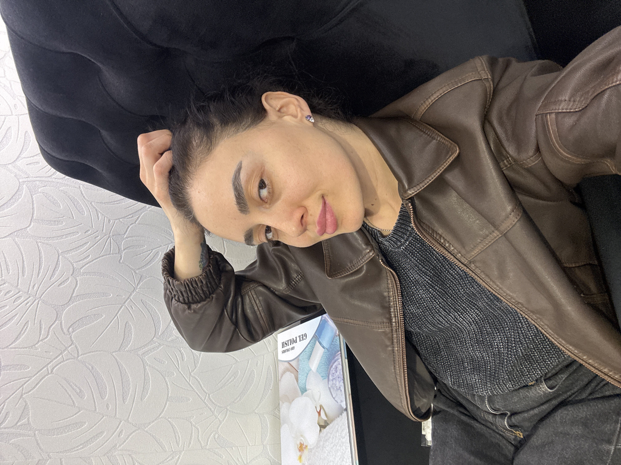 A woman wearing a brown leather jacket over a grey sweater and dark jeans is indoors, reclining against a black tufted sofa. She is resting her head on her hand. The wallpaper is white with a leafy pattern. A brochure or pamphlet with the words "GEL POLISH" and an image of orchids and towels is visible on the left. The location is likely Tbilisi, Georgia, based on details in the brochure that are partially visible.