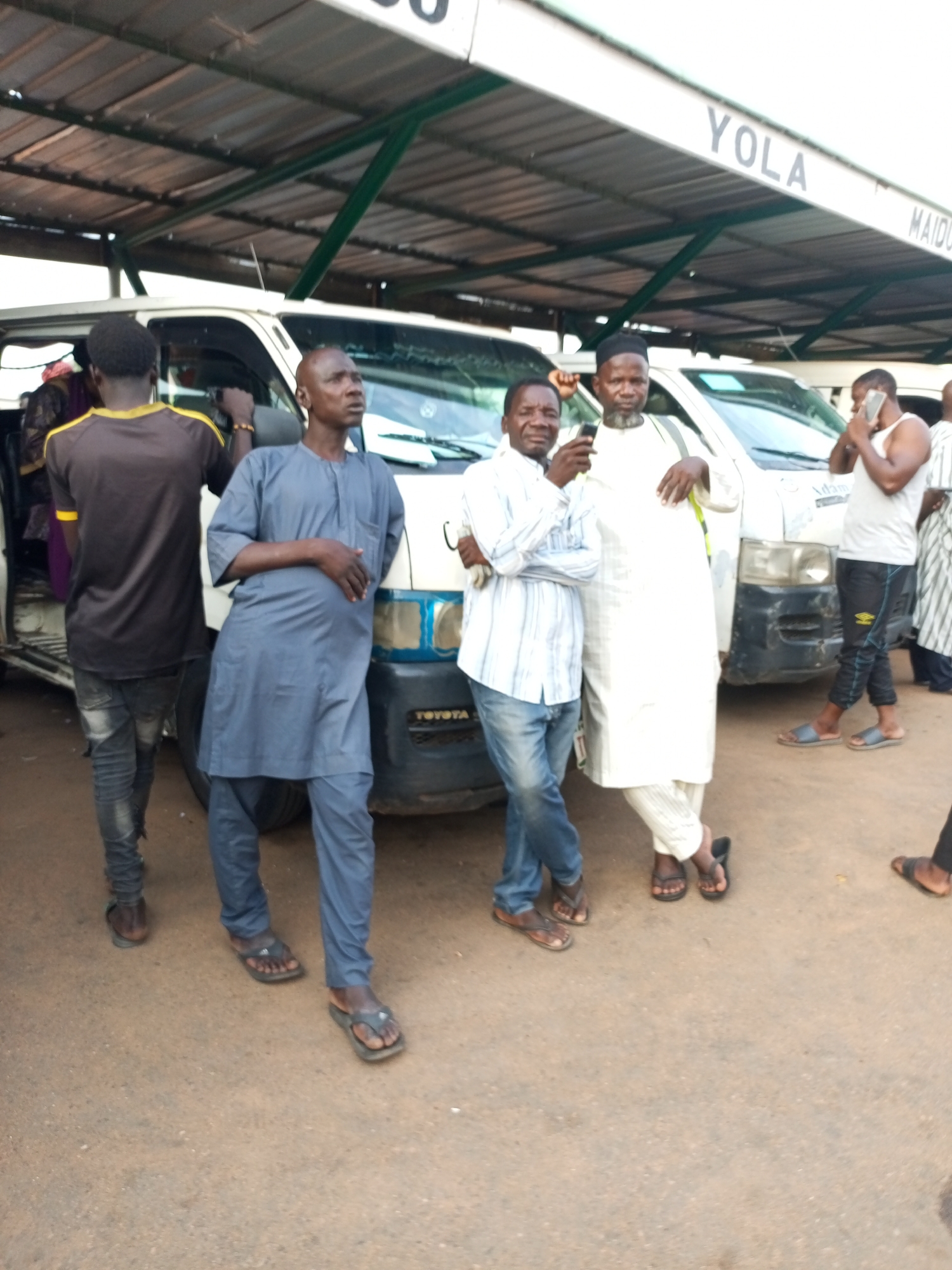 In Gombe, Nigeria, at a bus terminal with a covered waiting area, several men are gathered around a white Toyota Hiace minibus. The overhead structure, painted green and white, has the destination "YOLA" visible on a sign. The ground is a mixture of asphalt and dirt.
On the left, a man wearing a dark brown t-shirt with yellow trim and distressed jeans stands facing away from the camera. Next to him, in the center, a man in a flowing grey traditional outfit stands with his right hand resting on his stomach, looking upwards.
To the right of the man in grey, another man in a white traditional outfit with a matching cap is holding a mobile phone and looking at it. Beside him, a man in a white tank top and dark pants with yellow stripes, also holding a phone to his ear, stands with his legs apart. Another man, mostly out of frame on the far right, is wearing a striped garment. The minibus has a blue and white stripe on its front. The overall impression is a snapshot of everyday life at a transport hub.