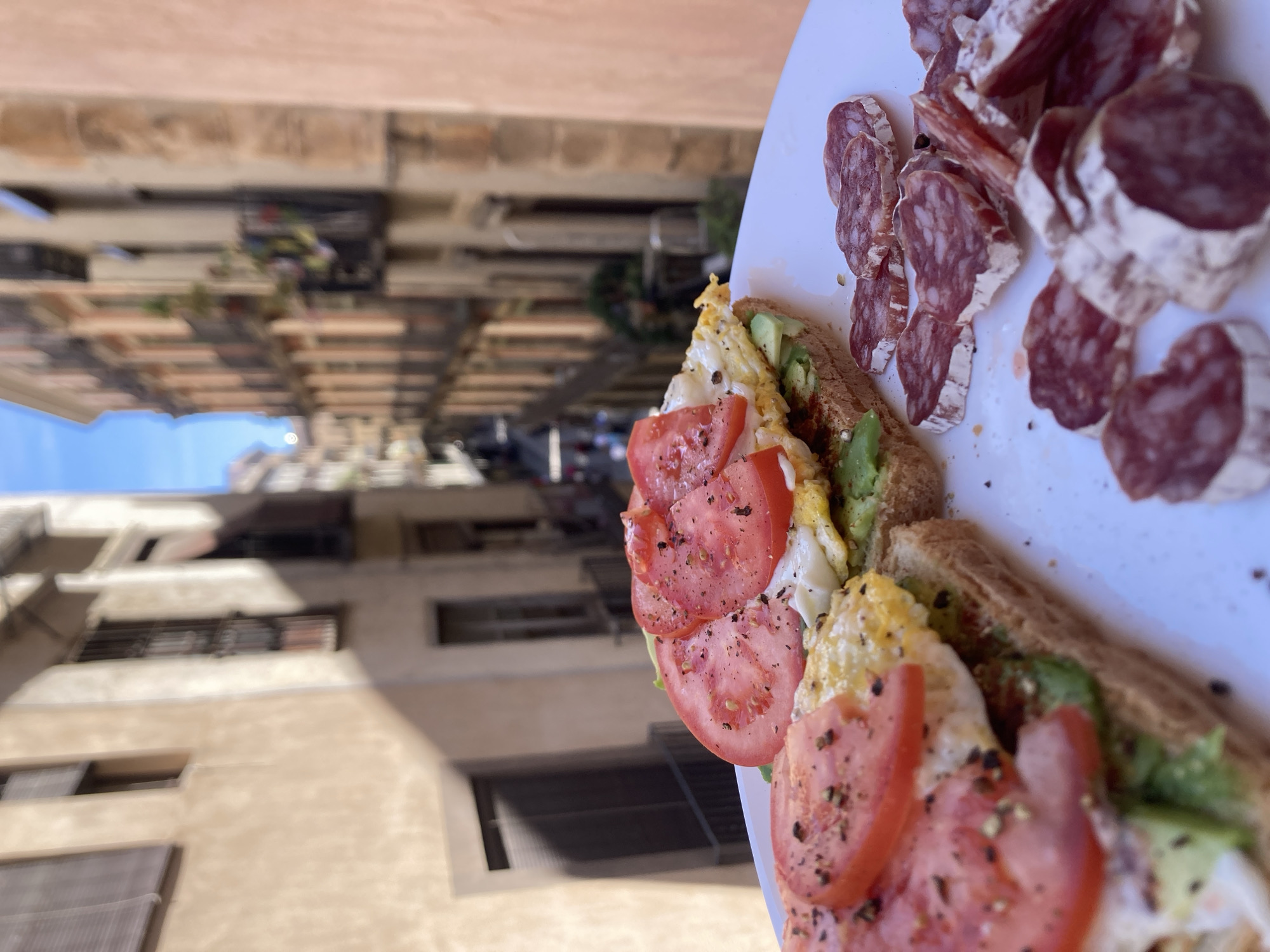 A medium shot shows an open-faced sandwich with tomato, avocado, and egg, next to sliced salami. The food sits on a white plate with a building exterior of Barcelona in the background. The building appears to be several stories high and has windows and balconies. The sky is blue, indicating that it is daytime. The focus of the image is on the food, with the background slightly blurred.