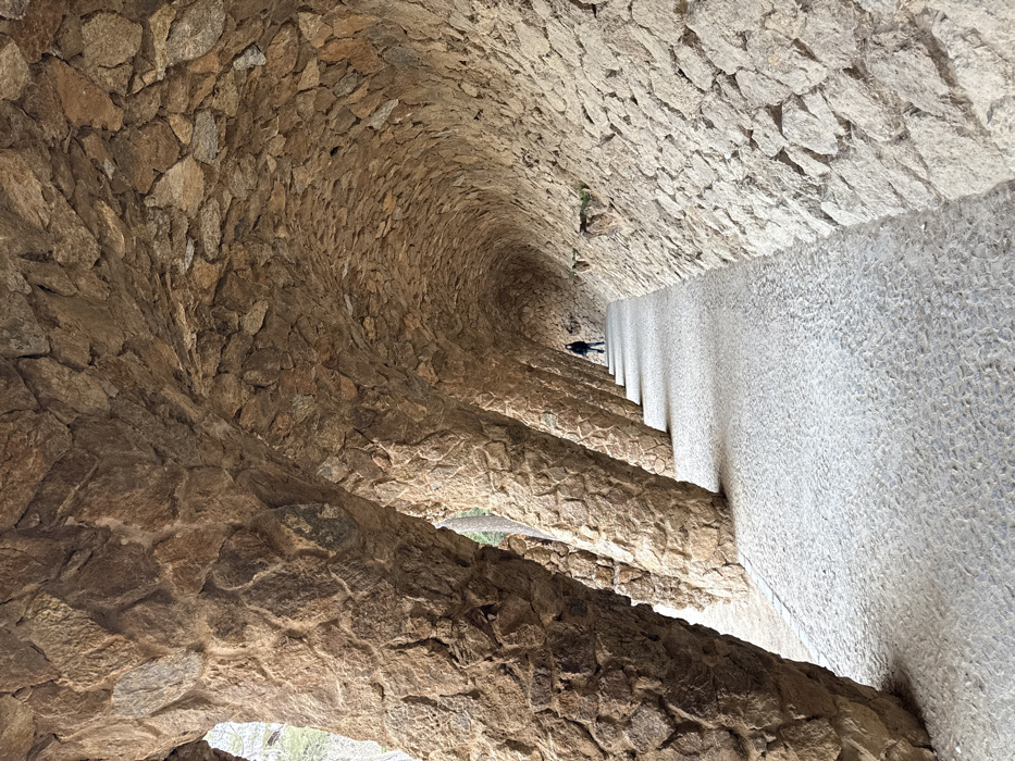 A view inside a stone-arched pathway at Park Güell, Barcelona, Spain. The scene features a long, curving walkway constructed from light-colored, irregularly shaped paving stones. On the left, a series of thick, textured columns, built from rough-hewn, light brown and grey stones, support an overhead archway. The ceiling, also composed of similar rough stones, curves organically to the right, forming a continuous, undulating wall that borders the right side of the path. Far in the distance, a lone individual in dark clothing is visible, standing or walking along the path. The entire structure is made of natural stone, illuminated by diffused light.