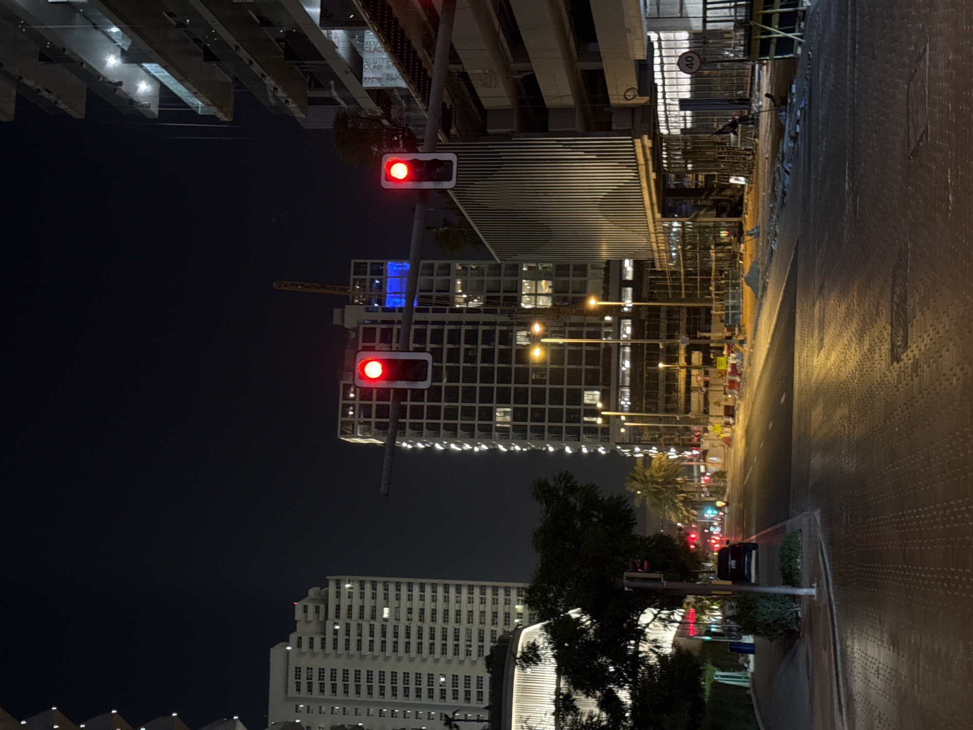 The image depicts a night scene in a city, likely Dubai, United Arab Emirates, given the architectural style and the presence of palm trees. The photograph is taken from a street level perspective, looking up towards illuminated buildings and a sky that is dark, indicating nighttime.
Dominating the foreground are traffic lights, with the top light showing a red signal, suggesting a stop. The traffic lights are mounted on poles, one of which is visible in detail. In the background, various buildings are visible. One prominent building features a grid-like facade with many illuminated windows. Another building, further in the distance, appears to be a multi-story structure with a repetitive window pattern. A construction site is also evident, with scaffolding and what appears to be a crane.
The street itself is paved and appears to be relatively empty of moving vehicles, though a dark-colored car is parked further down the road. The sidewalks are also visible, with some greenery, including trees and bushes, bordering them. The overall atmosphere is urban and nocturnal, with artificial lighting from buildings and streetlights creating a somewhat stark and modern aesthetic.
The image does not show any people clearly. There are no explicit weather cues, but the darkness suggests a clear night. There is a speed limit sign visible on a pole, showing "40" within a red circle, indicating a 40 km/h speed limit. The scene conveys a sense of a quiet urban environment at night, possibly in an area undergoing development or with modern architecture.