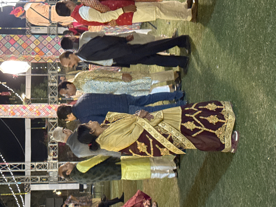 Several people stand outdoors, seemingly at an event. They are dressed in a mix of formal wear and traditional Indian attire. The background features decorative lighting and stage structures. A woman in a gold and maroon outfit stands out, with others dressed in suits or kurtas. The scene appears to be a social gathering.