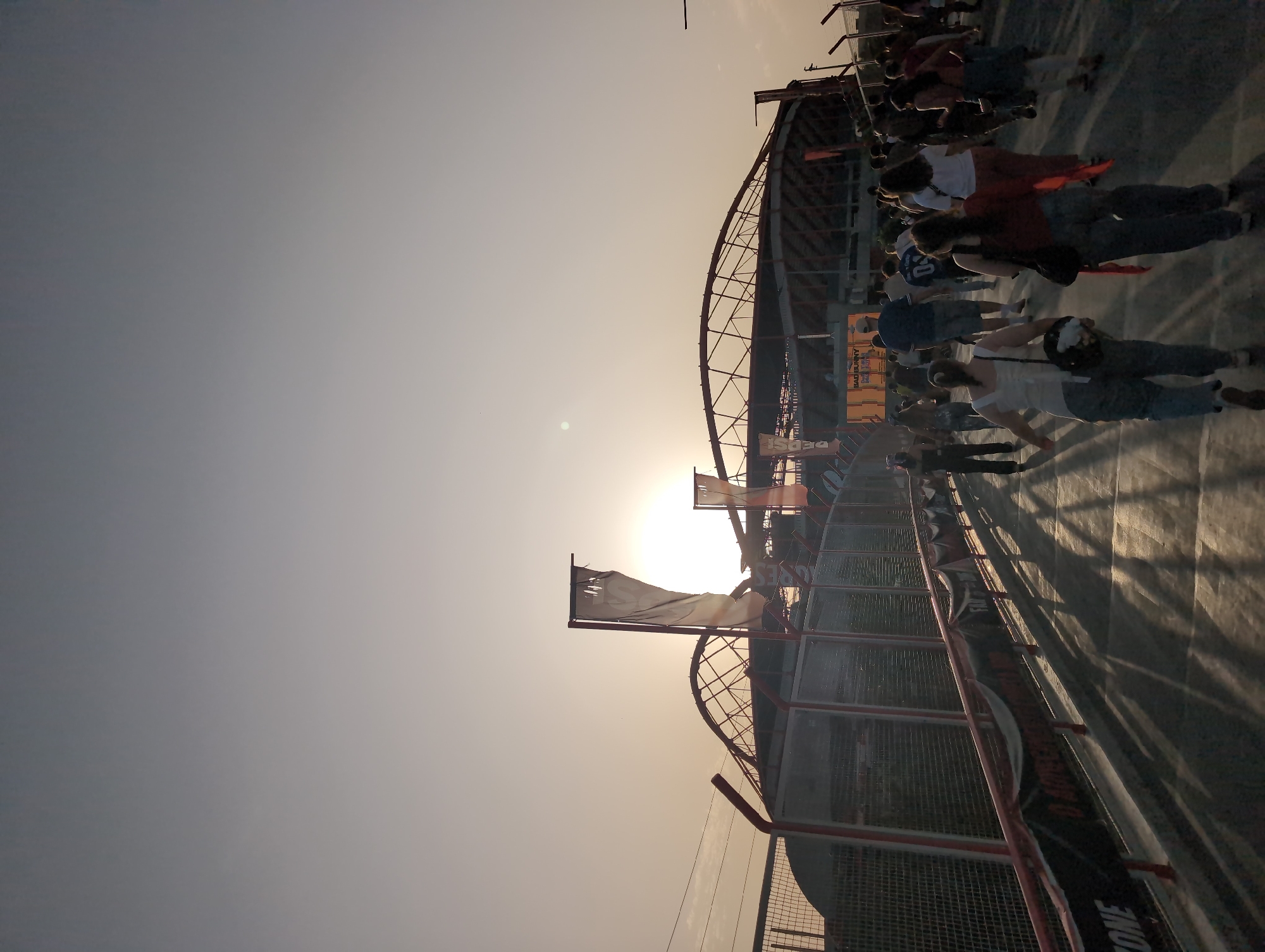 The image depicts a crowd of people at a stadium, likely located in Lisbon, Portugal. The sun is setting in the background, casting long shadows across the concrete floor. The crowd appears to be moving towards the stadium, presumably for an event. There are flags and banners visible, and the architectural structure of the stadium is visible in the background. The people appear to be casually dressed.
