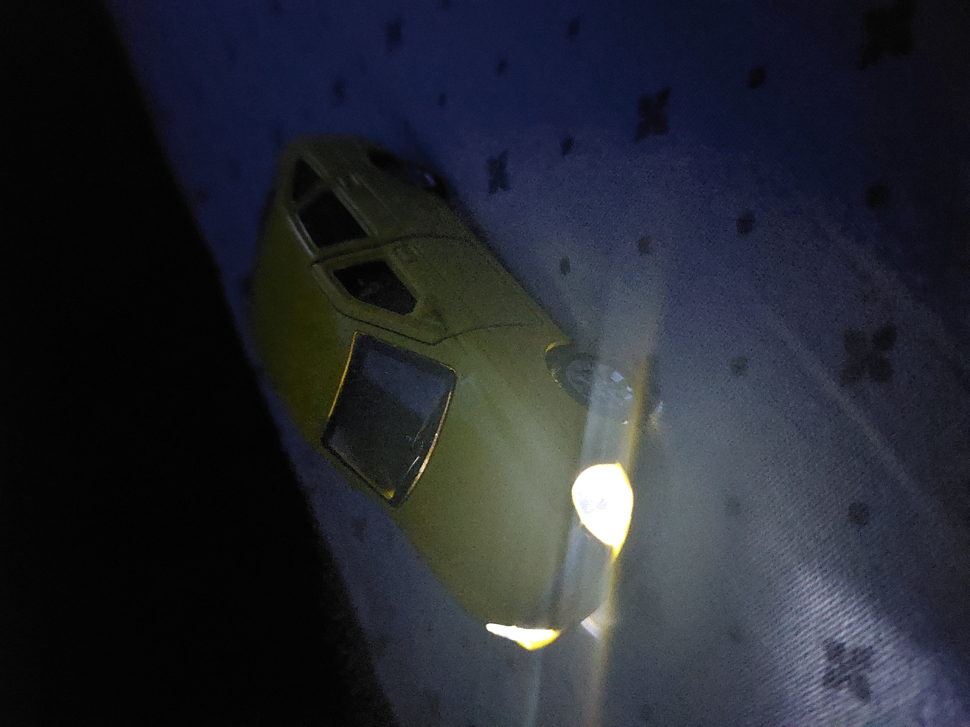 The image features a yellow toy car, likely a scale model, illuminated by its own bright headlights. The car is positioned diagonally on a dark blue or purple textured surface, which appears to be a fabric, possibly a blanket or carpet, adorned with faint, repeating dark patterns. The vibrant yellow light from the car's front headlights casts a strong glow downwards and forwards, serving as the dominant light source in the scene. A softer, yellowish glow is also perceptible from the rear of the vehicle. The overall dimness of the environment suggests the photograph was taken at night or in a very dark indoor space. The car appears stationary, with its lights simply switched on. No people or visible text are present in the image. While the location context is Chaikovsky, Russia, there are no specific visual cues within the photograph to indicate this.