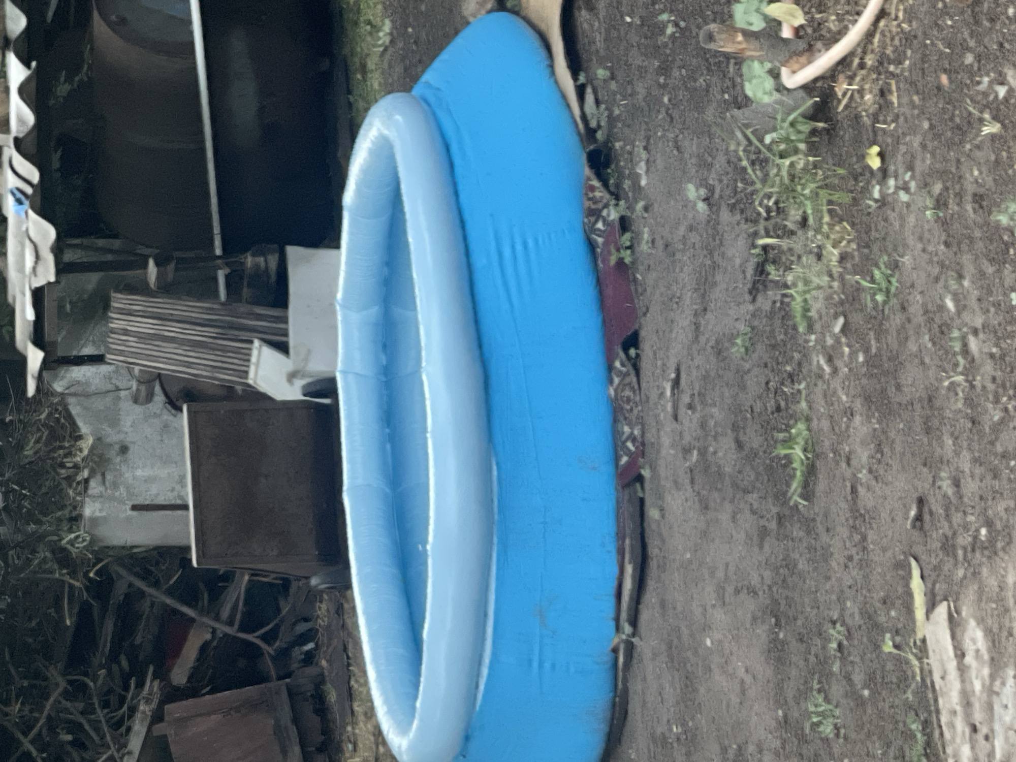 An inflatable, light blue kiddie pool is situated on the ground, partially filled with water. The pool is oriented vertically in the frame. To the left of the pool, there's a cluttered area with various items including what appears to be a dark-colored cylindrical object, possibly a tank or barrel, stacked wooden planks, and some metal or wooden crates. Some white fabric is also visible on the left side. The ground around the pool is predominantly dirt with sparse green vegetation growing in patches. A portion of a patterned rug or mat is visible beneath the pool. The overall scene suggests an outdoor, possibly backyard setting. The lighting indicates it is daytime. There is no visible text in the image. The image was taken in Bishkek City, Kyrgyzstan.