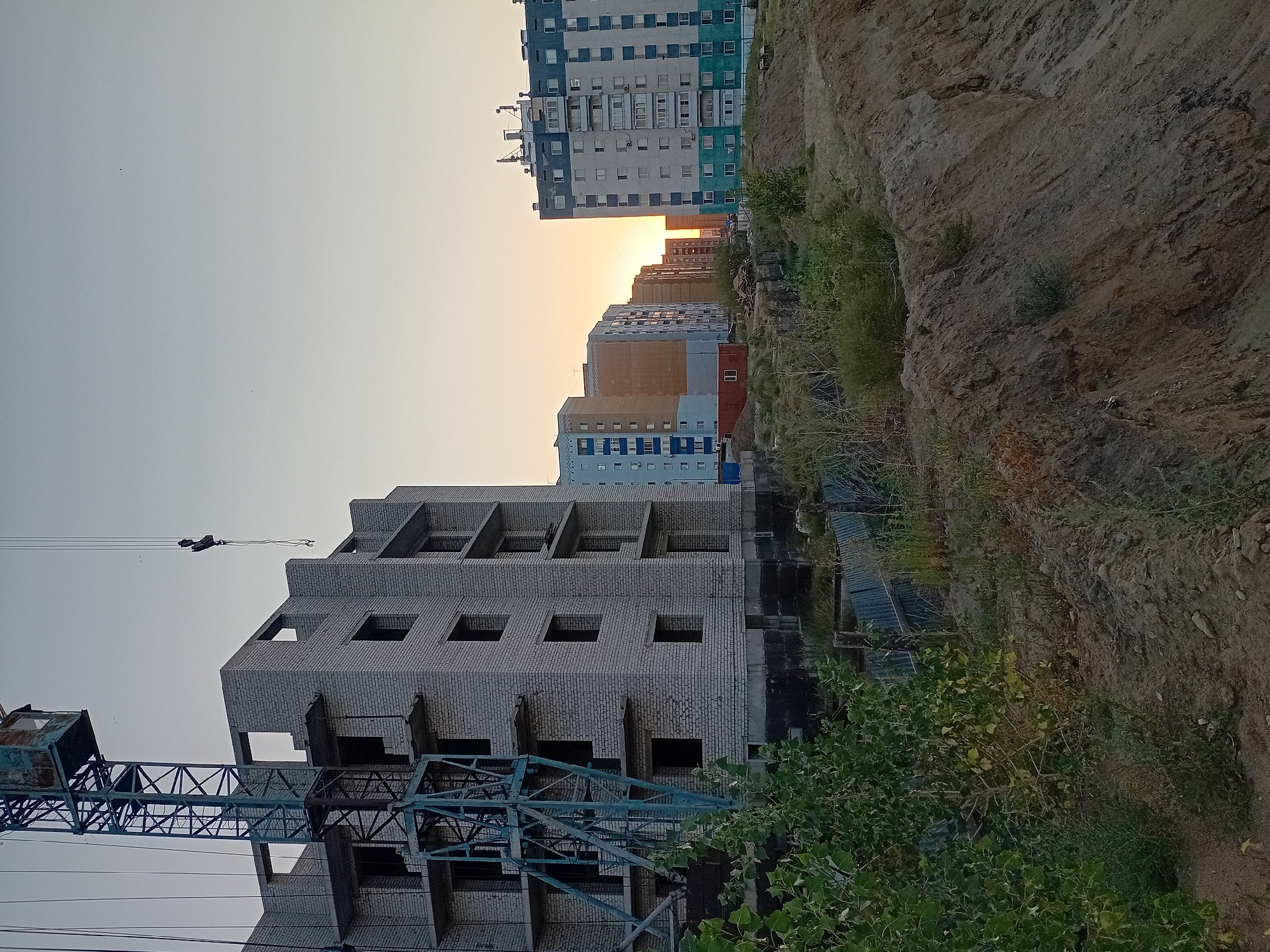 The image depicts a construction site in the city of Semey, Kazakhstan, during sunset. In the foreground, a large pile of dirt and vegetation suggests an ongoing excavation or landscaping project. To the left, a construction crane is partially visible, looming over an unfinished multi-story building constructed from light-colored bricks. The building has multiple floors with open window frames, indicating that it is still under construction.
In the middle ground, a row of completed residential buildings lines a street. These buildings vary in height and architectural style, with one prominent apartment complex featuring a blue and white facade. The setting sun casts a warm glow over the scene, illuminating the sky with hues of orange and yellow. The overall impression is one of urban development and progress in Semey. There are no people visible in the image.