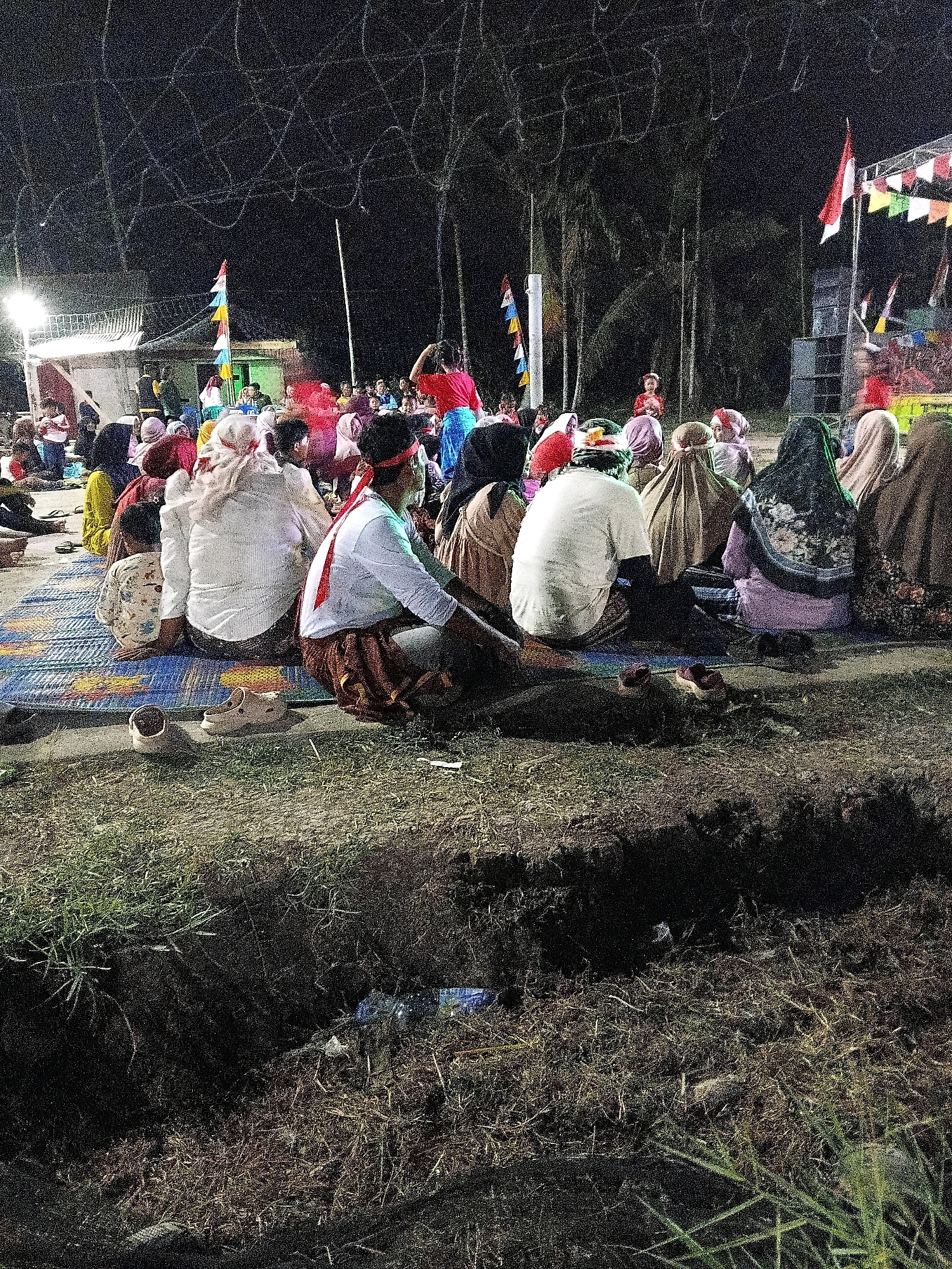 The image depicts an outdoor gathering at night in Margo Rukun, Indonesia, possibly a community event or performance. A crowd of people are seated on mats, facing what appears to be a stage or performance area in the background. There are decorative flags and banners strung across the scene, and some speaker equipment is visible. The lighting is dim, with artificial lights illuminating the central area and creating shadows. In the foreground, there is a dark, uneven patch of ground with some vegetation and debris, possibly a ditch or undeveloped area. The overall atmosphere suggests a festive or ceremonial occasion.