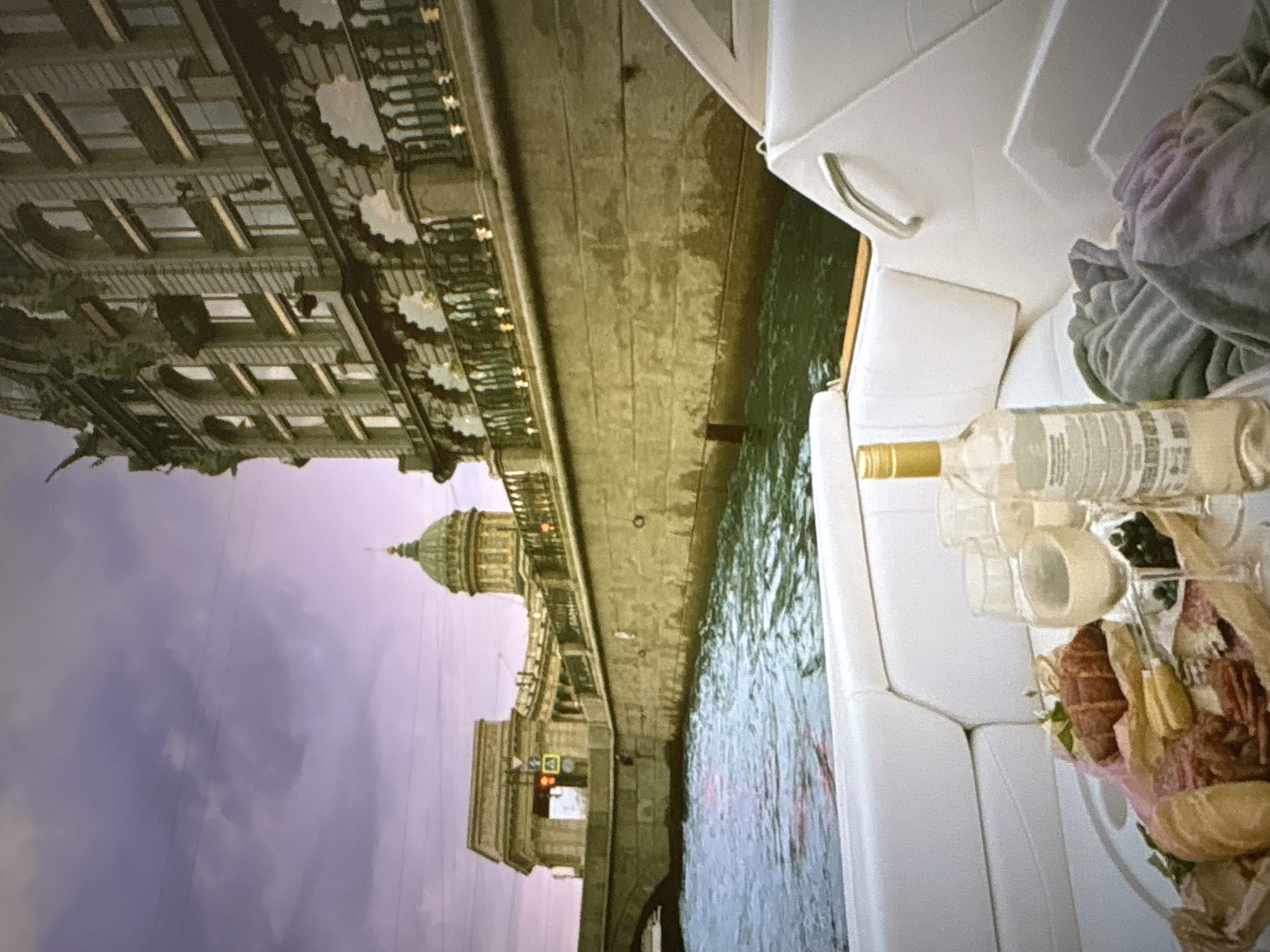 The image depicts a boat sailing on a canal, with the Kazan Cathedral and a neoclassical building on the right bank. The boat has white upholstered seating and a platter of charcuterie with bread, two glasses of white wine, and a bottle of white wine. A grey blanket is draped over the seats. The sky is a hazy purple and pink. The scene is set in St. Petersburg, Russia, not Pori, Finland.