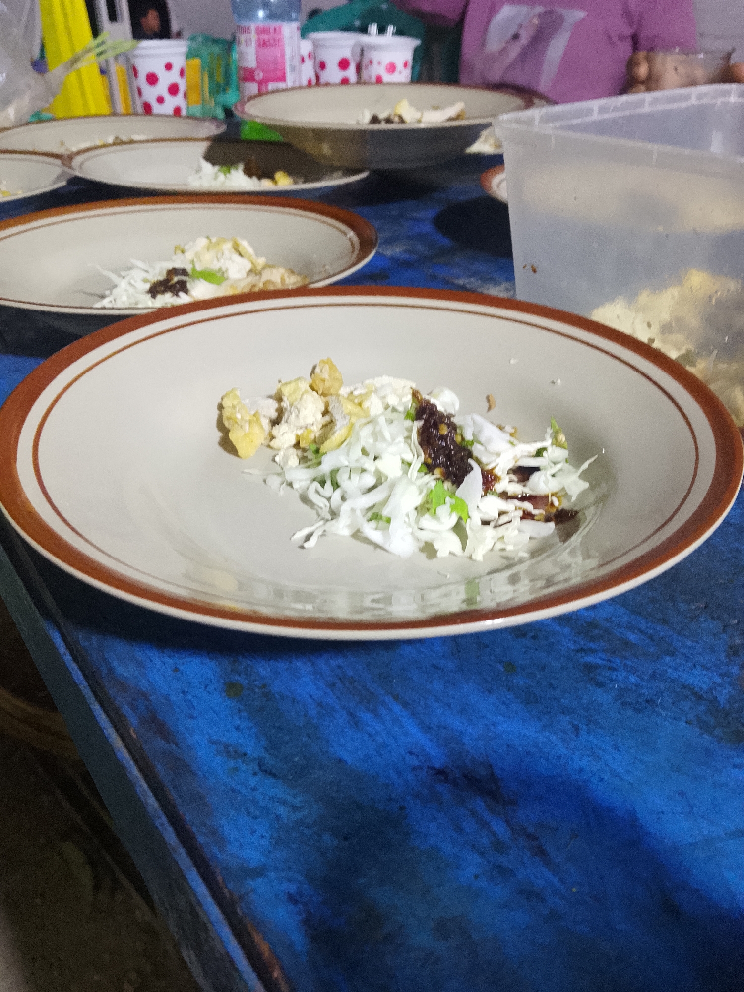 In Tanggungan, Indonesia, on a blue, weathered wooden table, a plate of food is centrally positioned. The plate, with a brown rim, contains shredded white cabbage, pieces of what appear to be fried tofu or tempeh, and a dark, possibly spicy sauce. The food is arranged in a casual, somewhat haphazard manner. In the background, out of focus, are other plates, plastic cups with red polka dots, and a translucent plastic container filled with food. A person in a pink shirt is partially visible in the upper right corner. The lighting suggests an evening setting.