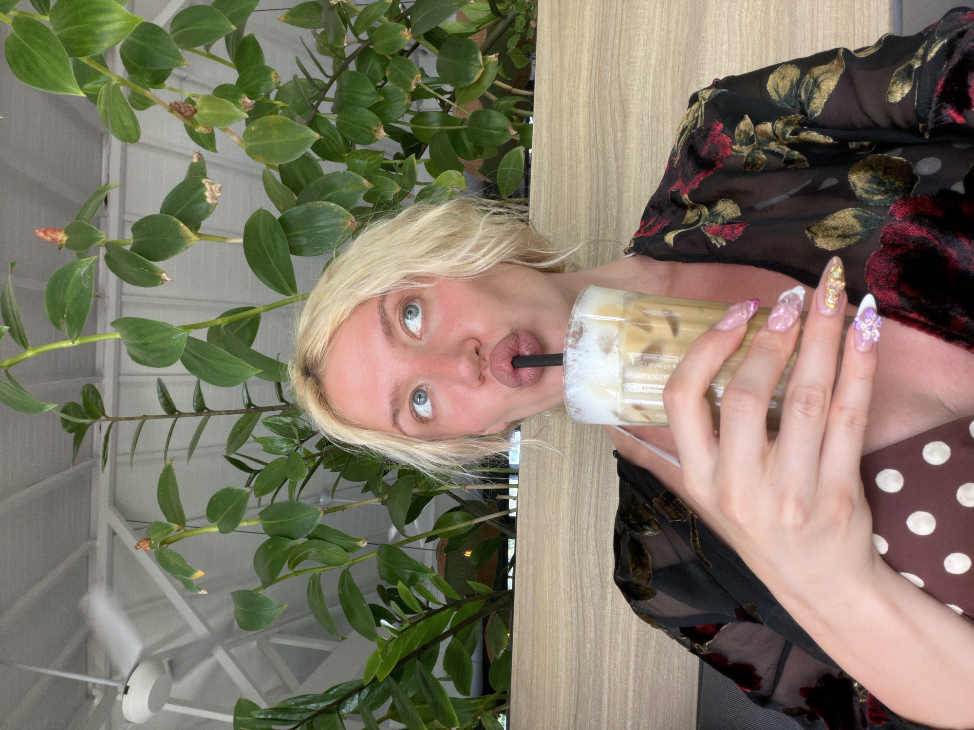 A blonde woman with light blue eyes is drinking an iced coffee through a black straw. She is wearing a sheer black top with a floral pattern in red and gold. Her nails are long and decorated with white, pink, and gold glitter, as well as small floral embellishments. She is seated in an indoor setting with a wooden partition behind her and lush green plants growing in front of a white ceiling. A white ceiling fan is visible in the upper left corner of the image. The overall setting suggests a cafe or restaurant in North Kuta, Indonesia.