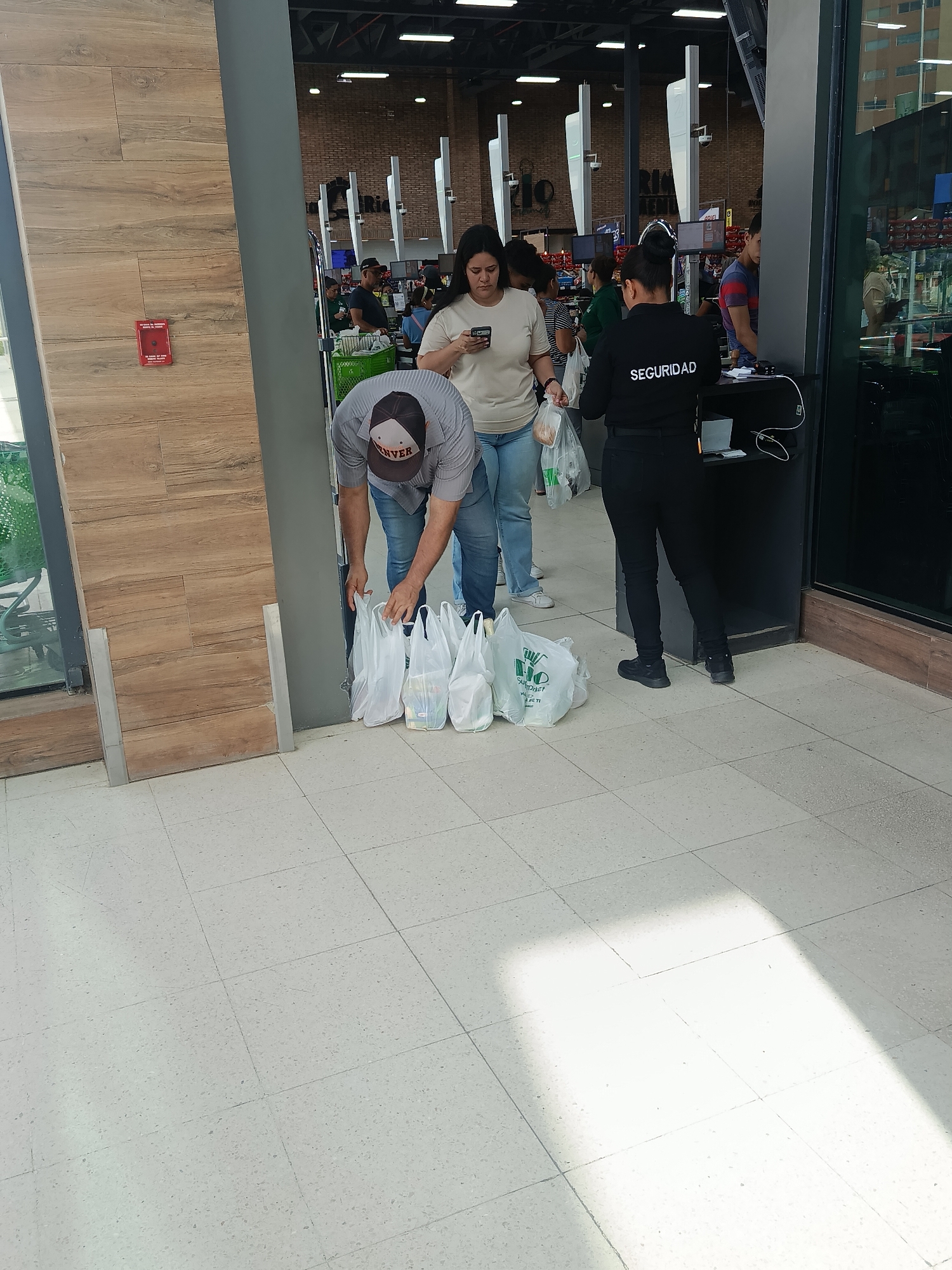 A wide shot depicts an indoor scene at what appears to be the exit or entrance area of a supermarket in Juan Griego, Venezuela, during daytime.
In the foreground, a man wearing a grey collared shirt and a baseball cap with "DENVER" printed on it is bending over, organizing multiple white plastic shopping bags on the light-colored tiled floor. Several bags are visible, some bearing the green "RIO" logo, indicating the supermarket's name. Beside him, a woman in a light-colored t-shirt and blue jeans is looking at her phone, holding another plastic shopping bag.
To their right, a security guard, identifiable by the "SEGURIDAD" text on her black uniform, stands facing inward, near a small black counter with some equipment, including a screen and charging cables. Behind these individuals, the interior of the supermarket is visible, showing checkout lanes, other shoppers, and shelves stocked with products. The background features exposed brick walls and various white structural elements, with more distant shopping aisles.
To the left, a tall, vertical wooden paneling section is prominent, with a red fire alarm pull station mounted on it. The floor is light-colored tiled with visible grout lines, and a bright patch of sunlight reflects on it towards the lower right. The overall atmosphere is routine, with people engaged in their shopping-related activities.