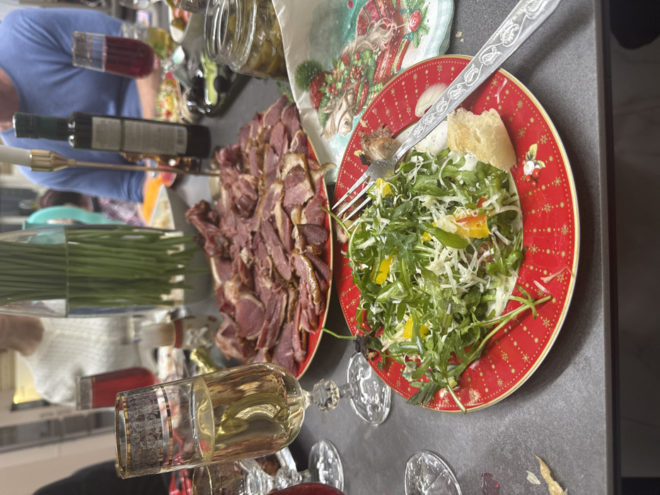 A countertop features platters of food: one with sliced meat and another with a salad of greens, shredded cheese, and colorful peppers, alongside a slice of bread. A silver fork rests on the salad plate. There are beverages, including a glass filled with white wine and a bottle of olive oil. The scene includes glassware, possibly wine glasses, and fresh green onions. A festive napkin with a Christmas motif is also present.