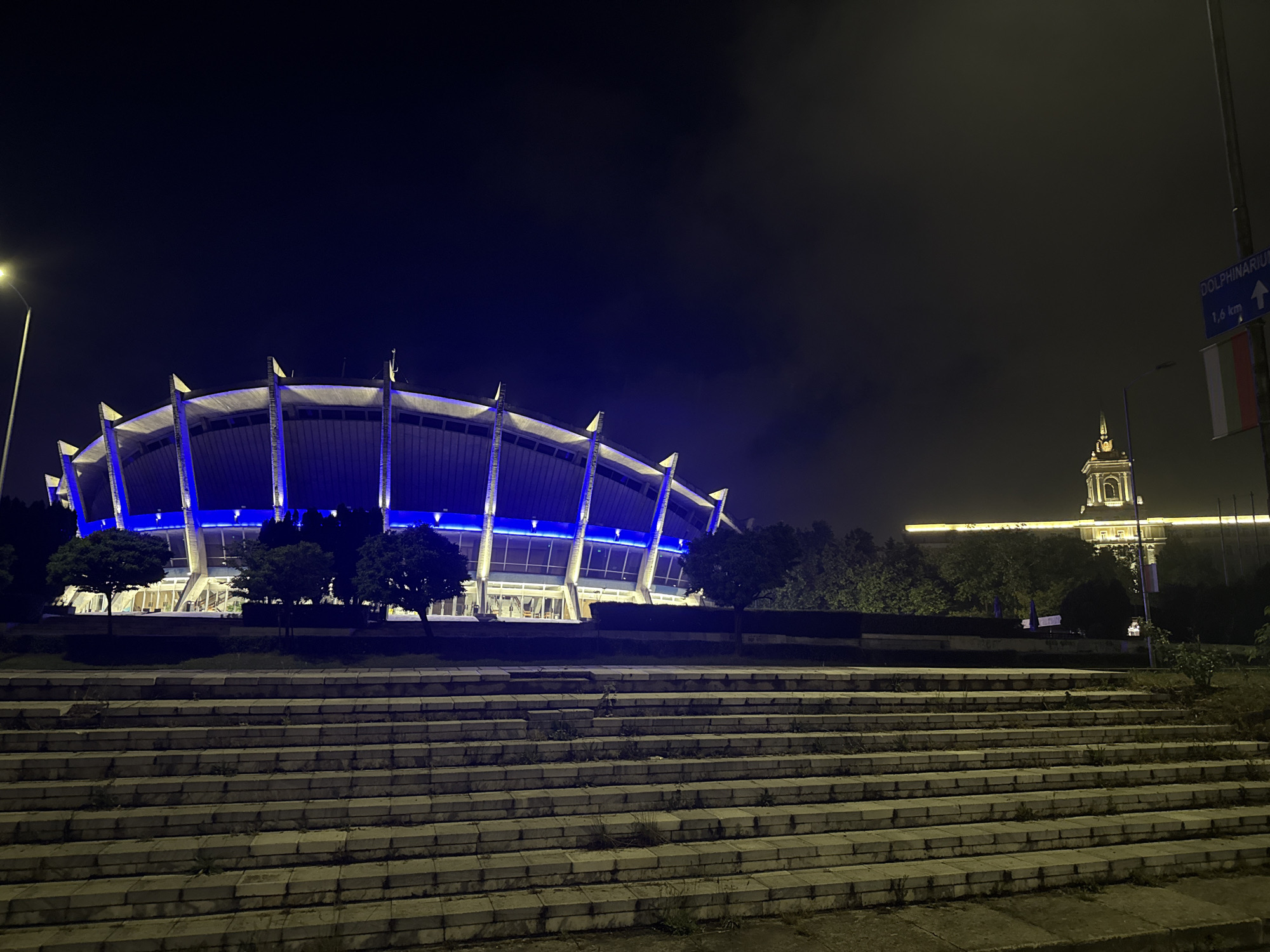 The image presents an illuminated urban night scene featuring distinctive architectural structures. In the left and central portion, a large, modern building, possibly an arena or stadium, is brightly lit with vibrant blue lights accentuating its circular or elliptical facade and ribbed roofline. Several trees are positioned around its base.
To the right and further in the background, a classical-style building with a prominent, warmly lit clock tower or turret stands out against the dark sky. This structure contrasts in design and illumination with the blue-lit building in the foreground.
The immediate foreground features a broad set of concrete or stone steps, appearing weathered with grass and weeds growing in the crevices. A streetlamp on the far left emits a yellow glow. The sky is dark, characteristic of nighttime, with some areas showing a subtle haze.
On the far right, a street pole holds a blue directional sign that reads "DOLPHINARIUM" and "1,6 km," accompanied by an upward-pointing arrow. Below this sign, a flag is visible, displaying horizontal stripes of white, green, and red, which identifies it as the national flag of Bulgaria. The scene depicts a quiet nocturnal view of landmarks in what appears to be a Bulgarian city.