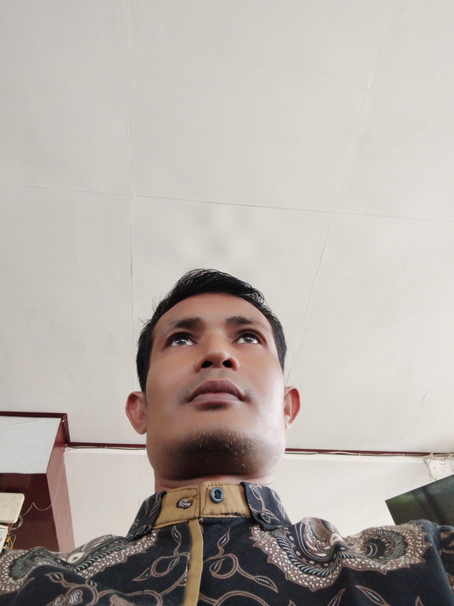 A low-angle, medium close-up shot captures an Indonesian man gazing intently upwards and slightly to the right. He has dark hair, dark eyes, and noticeable stubble on his chin and jawline. His facial expression is neutral, appearing contemplative or observant of something above the camera's view.
The man is wearing a dark shirt, likely a batik print, characterized by intricate brown and cream patterns resembling foliage or abstract motifs. The shirt has a distinctive light brown collar and placket with dark buttons.
The background is dominated by a plain, light-colored (likely white or off-white) ceiling, which occupies more than half of the frame. The ceiling shows faint lines and seams, suggesting it might be made of panels. On the left side of the image, a hint of a dark brown or maroon structural element, possibly a beam or part of a wall, is visible. On the far right, a dark, indistinct shape can be seen, possibly part of another object in the room.
The setting is indoors, with soft, ambient lighting that evenly illuminates the man's face, suggesting either overhead artificial lights or indirect natural light. There are no visible clues to indicate the specific time of day or outdoor weather conditions. The man's attire, particularly the patterned shirt, is culturally consistent with the provided location context of Lhokseumawe, Indonesia. There is no visible text in the image.