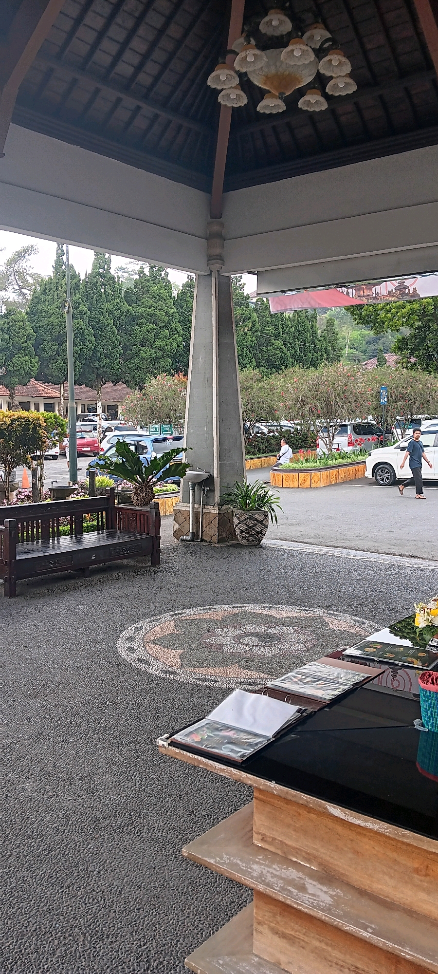 In Baturiti, Indonesia, an outdoor scene unfolds beneath a covered walkway with a wooden-tiled roof and a multi-light chandelier. A concrete pillar supports the roof. In the foreground, a dark wooden bench sits on a textured gray pavement adorned with a circular mosaic pattern. To the right, a wooden table displays open menus with glossy pages. Beyond the walkway, a parking lot is visible, with various cars including a white SUV and a red sedan. Lush green trees, some with red flowers, line the background, and distant buildings are partially obscured. Two individuals are present; one man walks towards the right side of the frame, and another person, partially visible, stands in the parking lot. A potted plant with large green leaves is situated near the concrete pillar, and another potted plant with woven texture sits to its right.