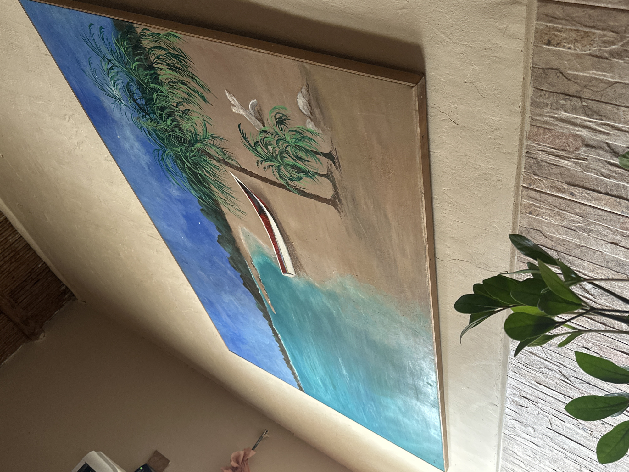A painting of a tropical beach scene hangs on a textured, light beige wall. The painting depicts a turquoise ocean meeting a sandy beach, with a red and white boat resting on the shore. Palm trees are depicted on the beach, and a distant coastline is visible in the background under a blue sky. The painting is framed with a light brown wooden border. In the foreground, a potted plant with dark green leaves is visible against a textured wall with vertical stonework. The overall scene suggests an indoor setting, possibly a home or restaurant, in El Maco, Venezuela.