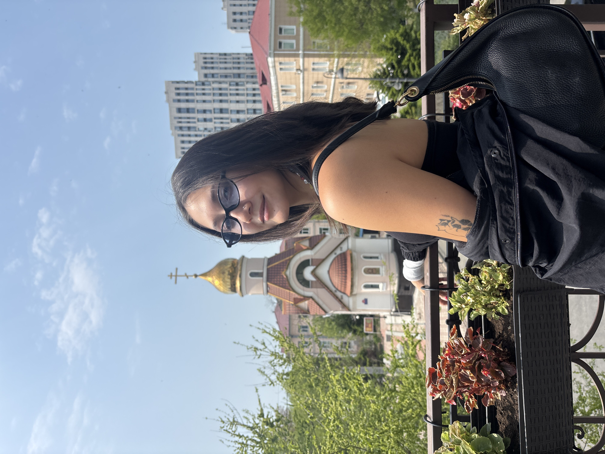 A slightly rotated medium shot features a woman wearing eyeglasses, with a tan-colored arm visible, posing against an urban backdrop. Behind her, there's a golden-domed Russian Orthodox church, trees, and high-rise apartment buildings. A black jacket and black leather purse are visible on her right side. The woman looks towards the camera. The scene seems to be taken during the daytime. The background buildings and church dome indicate the location is likely Vladivostok, Russia.