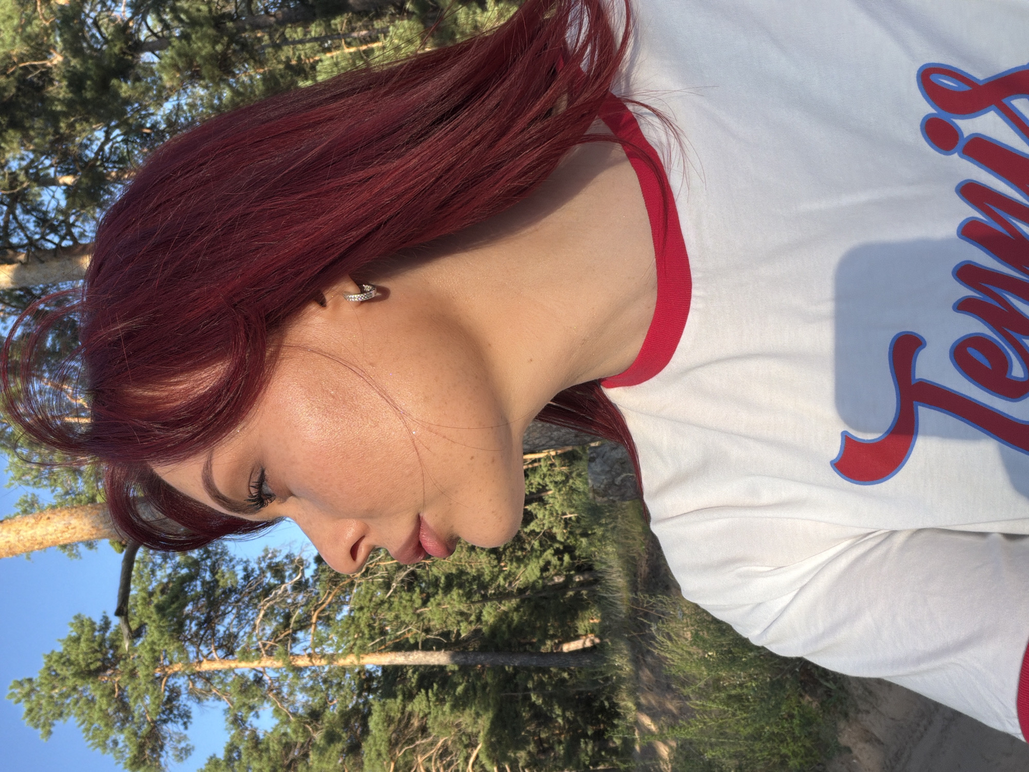 The image shows a medium shot of a young woman with dark red hair, seen from the side, looking to the left. She has fair skin with freckles and is wearing a white t-shirt with red trim and blue and red lettering that appears to spell out "Tennis." She has a small hoop earring in her left ear. The background consists of a forest with tall pine trees and a clear blue sky. The lighting suggests it is either early morning or late afternoon, with sunlight illuminating the woman's face and the trees. The location is identified as Uzukovo, Russia.