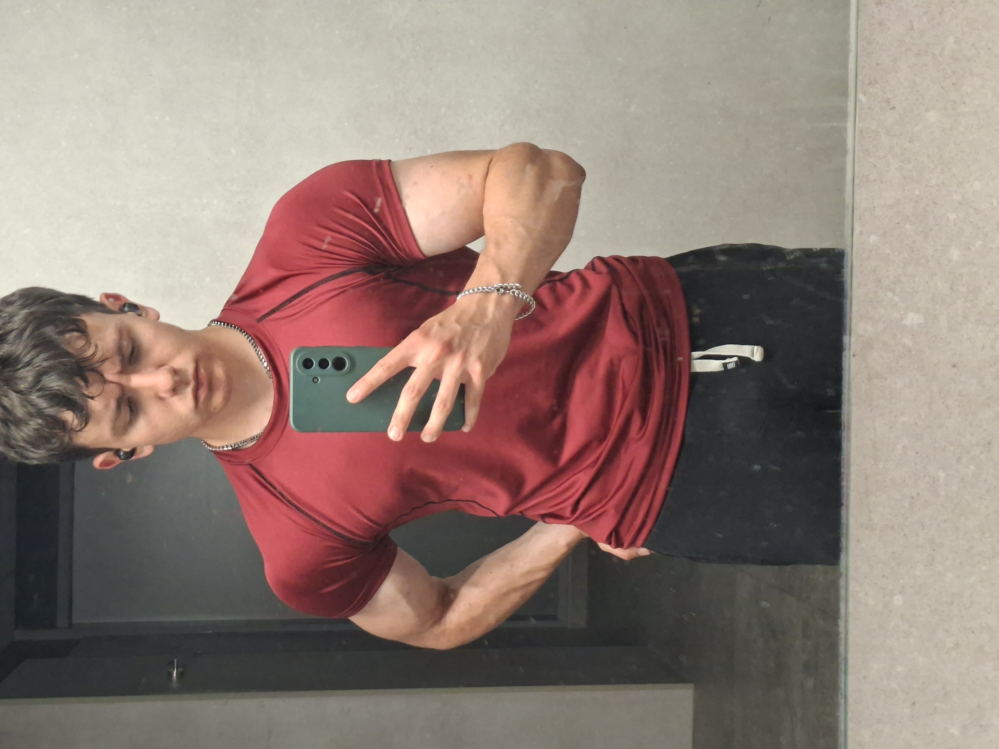 A young man takes a mirror selfie in what appears to be an indoor setting, possibly a gym changing room or bathroom, in Plovdiv, Bulgaria. The man, with dark, curly hair, is wearing a dark red athletic t-shirt with black detailing and black shorts or sweatpants. He is accessorized with a silver chain necklace, a silver beaded bracelet on his right wrist, and black earbuds in both ears. He holds a smartphone with a distinct green case and visible triple camera lenses to capture the reflection.
The man appears to be flexing his right arm, showcasing well-defined biceps and forearms with visible veins. His left arm is also slightly flexed. He is looking down at his phone or at his reflection, with his eyes partially closed, seemingly focused on the pose or the photograph.
The background visible in the mirror shows a dark grey or black surface, likely a countertop or floor. Beyond the mirror, a light grey or beige tiled or textured wall is visible. On the white drawstring loop of his shorts, there is some small, indistinct black text. The overall scene suggests a moment of personal fitness documentation or self-appraisal.