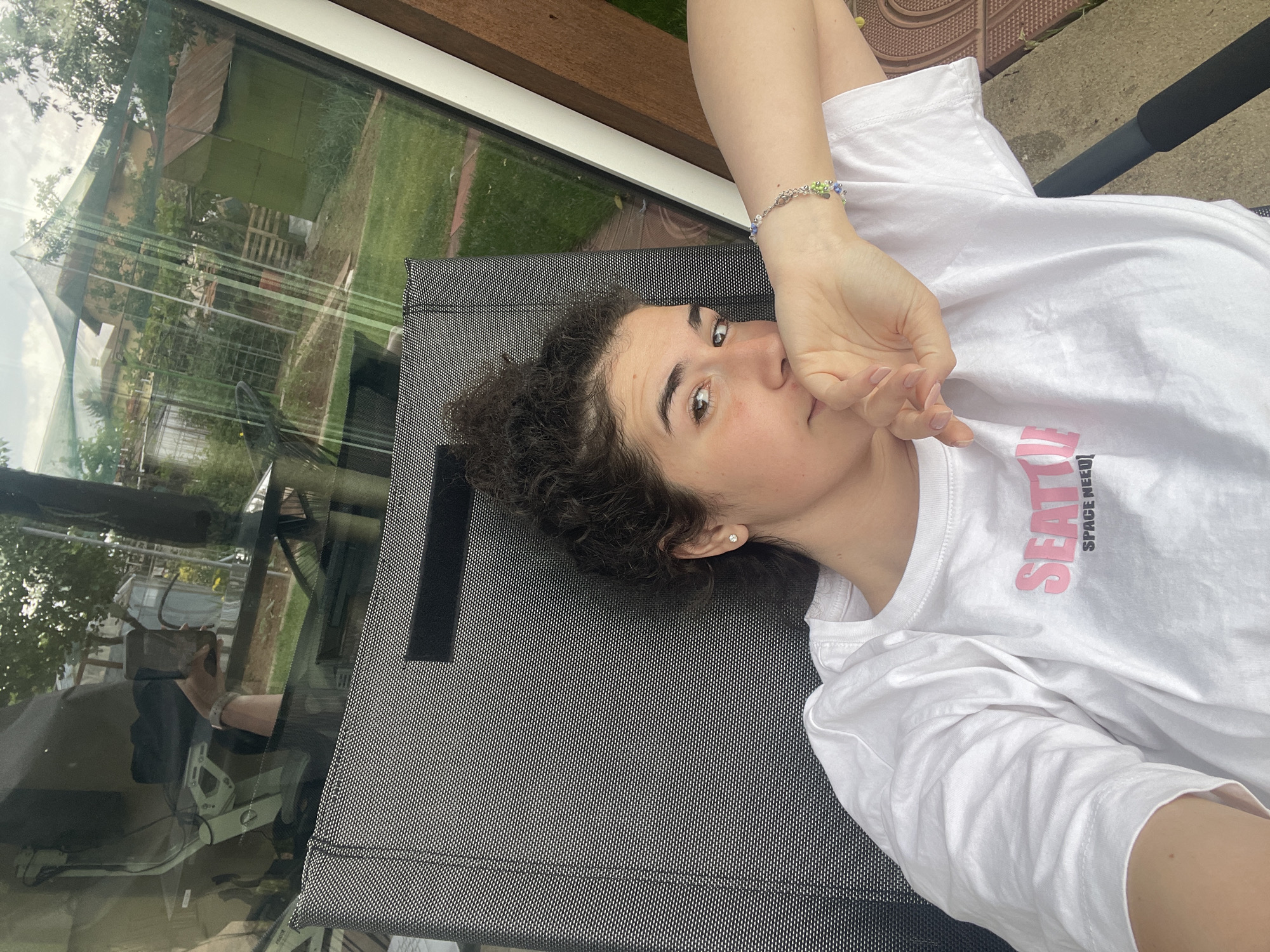 A fair-skinned woman with curly dark hair and an earring in her left ear is lying on a black mesh-like surface. She is wearing a white t-shirt with a pink inscription "SEATTLE SPACE NEEDLE". She is holding her right hand near her face, wearing a bracelet with blue and green beads on her wrist. A window reflects a view of a green garden, trees, and buildings. The woman appears to be taking a selfie. The location is Sofia, Bulgaria.