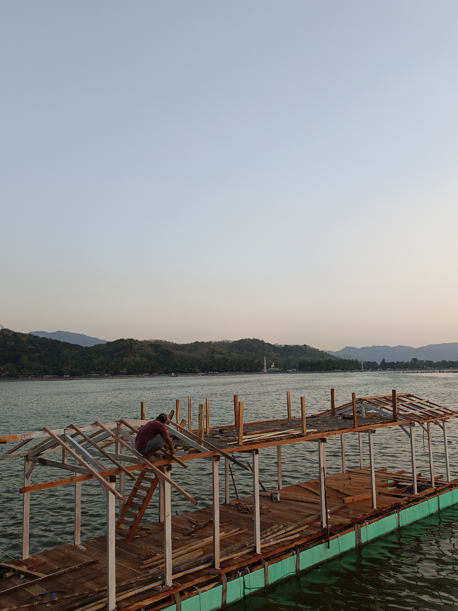The image shows a man working on the roof of a wooden structure on a body of water, possibly Krakitan, Indonesia. The man, wearing a maroon shirt and blue shorts, is crouched on a ladder, appearing to be involved in construction or repair. The structure is unfinished, with exposed wooden beams and planks scattered on the platform. The platform itself appears to be floating, supported by green pontoons. In the background, a lush green hillside with trees stretches across the horizon, and distant mountains are visible. The water is calm, reflecting the light blue sky. A white dome, possibly a building, is visible on the distant shoreline. The scene suggests a rural or coastal setting.