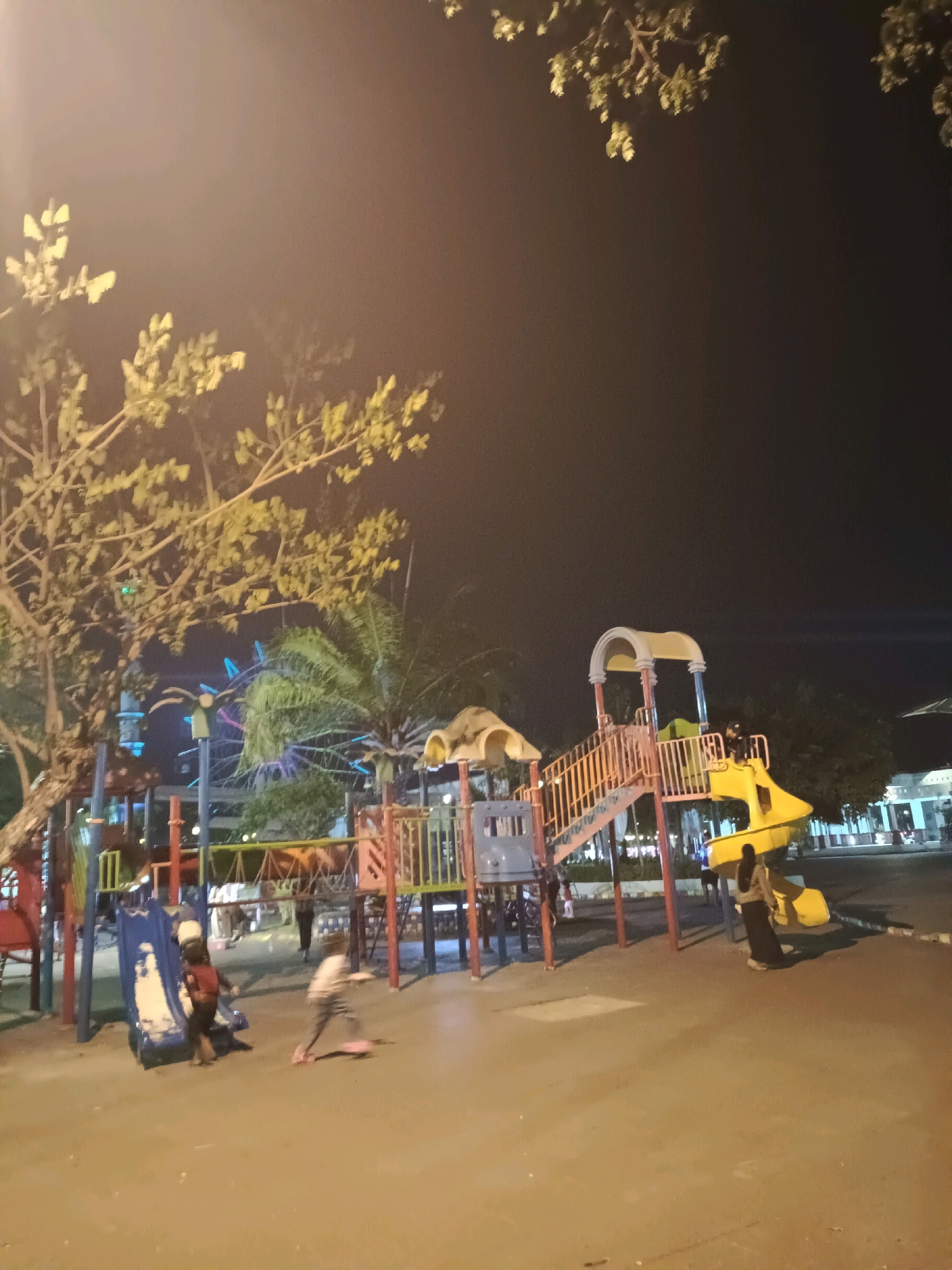 A nighttime scene at a playground in Lamongan, Indonesia, features a colorful, multi-level play structure with slides and stairs. Several children are present. One child is on a blue slide, while another is on a pink scooter. An adult, possibly a parent, is visible near the play structure. Trees with foliage frame the upper part of the image, and a Ferris wheel is visible in the background. The ground is paved, and streetlights illuminate the area.