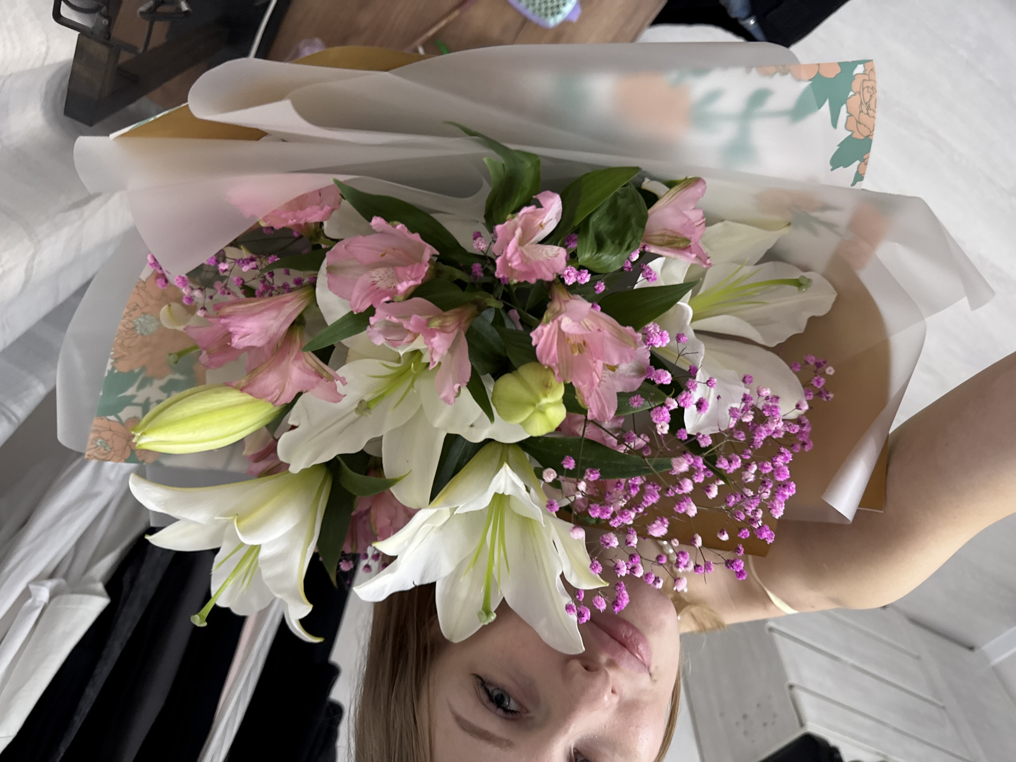 The image shows a young woman holding a bouquet of white lilies and pink alstroemerias. She is in an indoor setting, likely a bedroom, with a white comforter visible. A dark wooden object is present in the background. The woman is looking directly at the camera with a neutral expression. The bouquet is wrapped in translucent paper with a floral pattern. The lighting suggests it is daytime, but there are no clear indicators of the weather or specific time of day. The image appears to be a selfie, given the perspective. The location is identified as Perm, Russia, though no specific visual cues in the image confirm this.