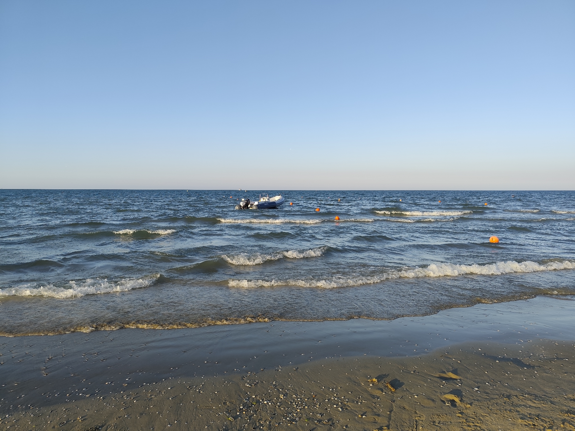 This is a wide shot of a beach in Rimini, Italy, under a clear blue sky. Gentle waves lap onto the sandy shore, which is scattered with small shells. In the distance, a small white boat with two outboard motors is moored. Several bright orange buoys are also visible in the water, marking an area offshore. The overall scene depicts a peaceful coastal environment.