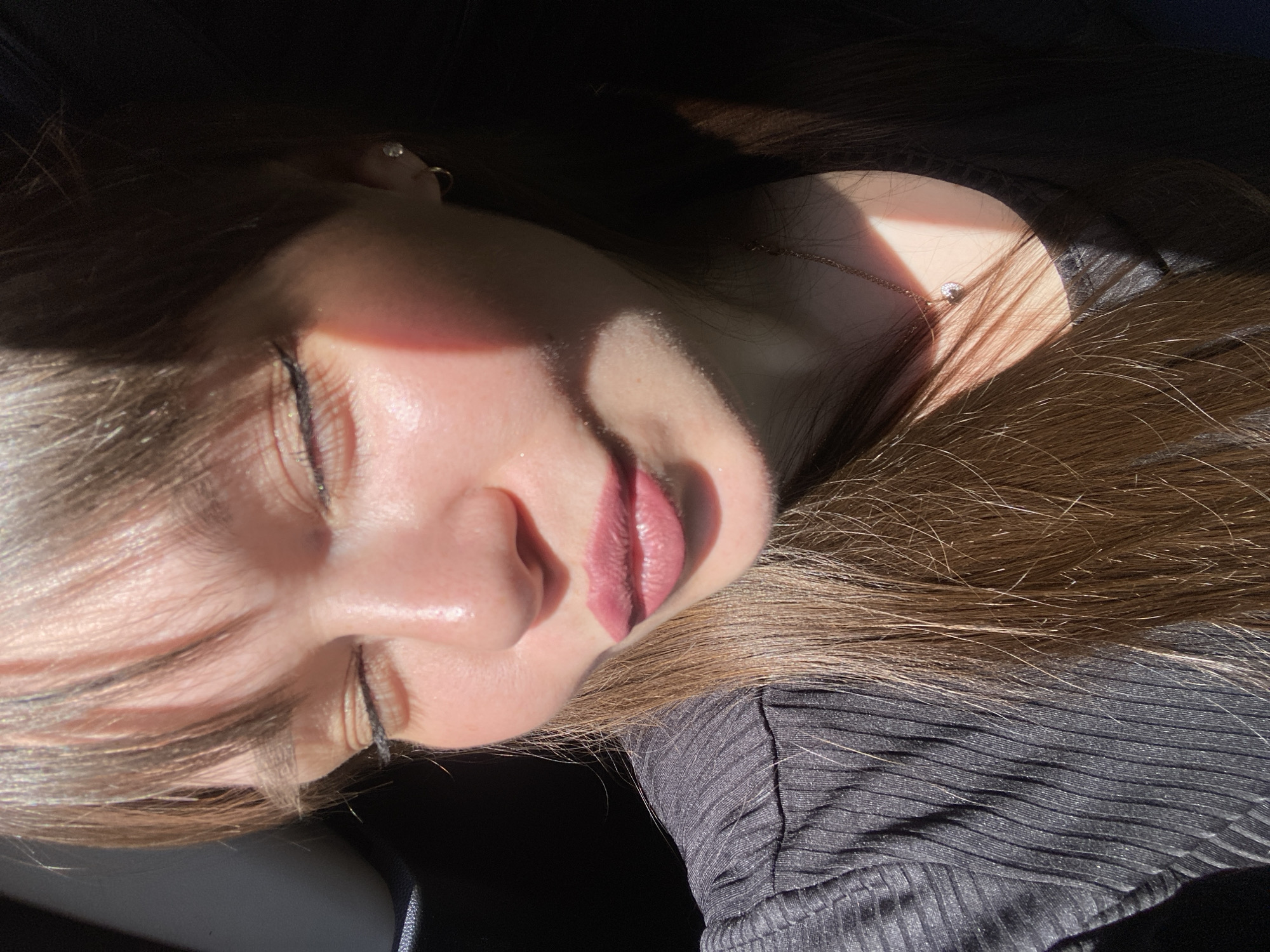 A close-up, slightly rotated image shows a young woman with closed eyes, her face illuminated by sunlight. She is lying down, likely indoors. Her hair, brown with light ends, is spread around her face and neck. She has fair skin and is wearing makeup, including lipstick and eyeliner. A small piece of jewelry is visible on her neck. She is wearing a black, textured top. The location in the city of Votkinsk, Russia is not indicated in the image.