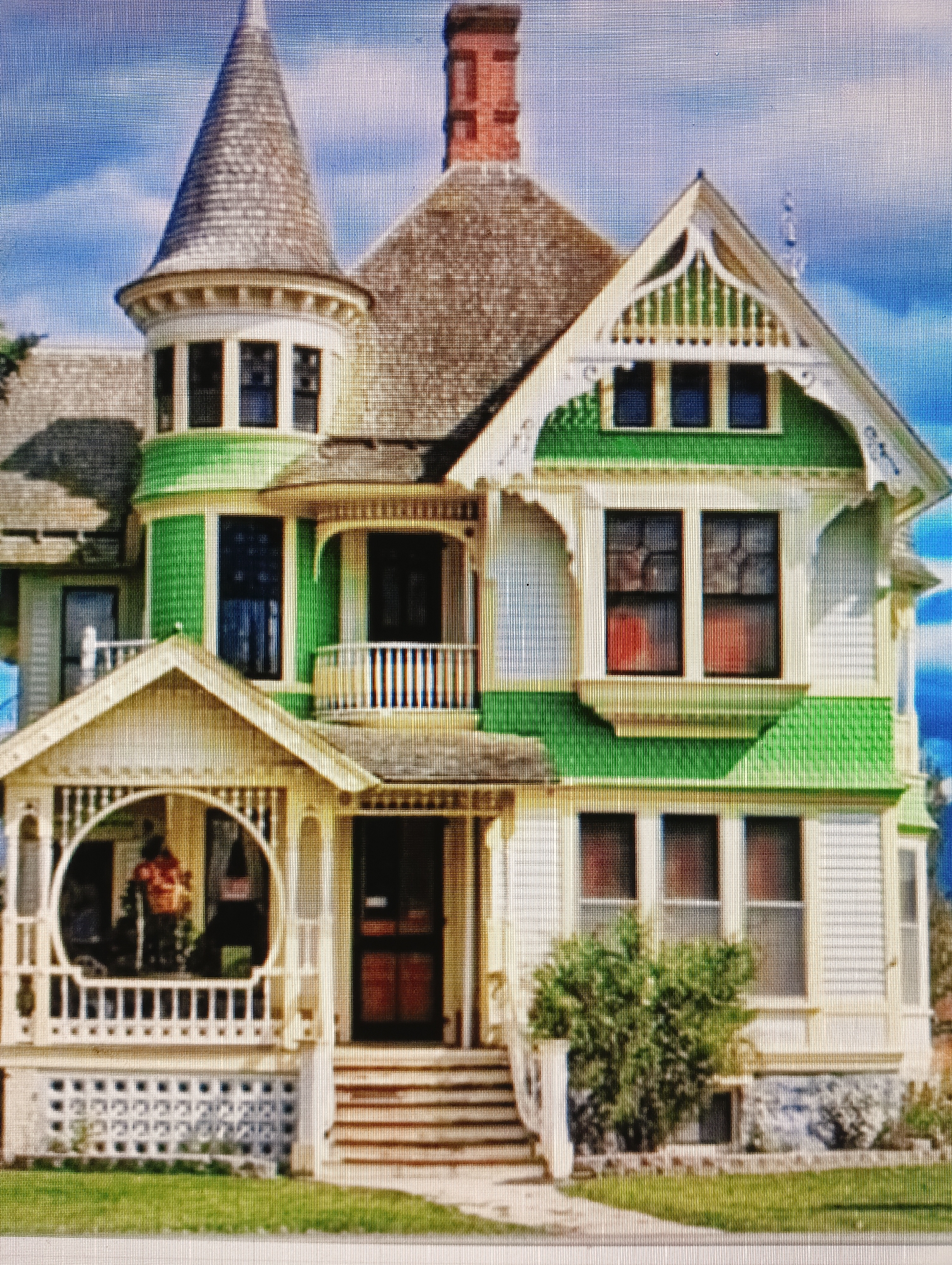 The image is a photograph of a large, multi-story house with a distinctive architectural style. The house is primarily white with vibrant green accents. It features a conical turret with a shingled roof, multiple windows of various sizes and shapes, and a prominent front porch with a decorative railing and a rounded opening. A brick chimney rises from the roof. The house is surrounded by a lawn and appears to be in a suburban or rural setting, with a blue sky and scattered clouds visible in the background. There are no people visible in the image. The provided image does not contain any visual information that would allow for its location to be identified as Sluck, Belarus.