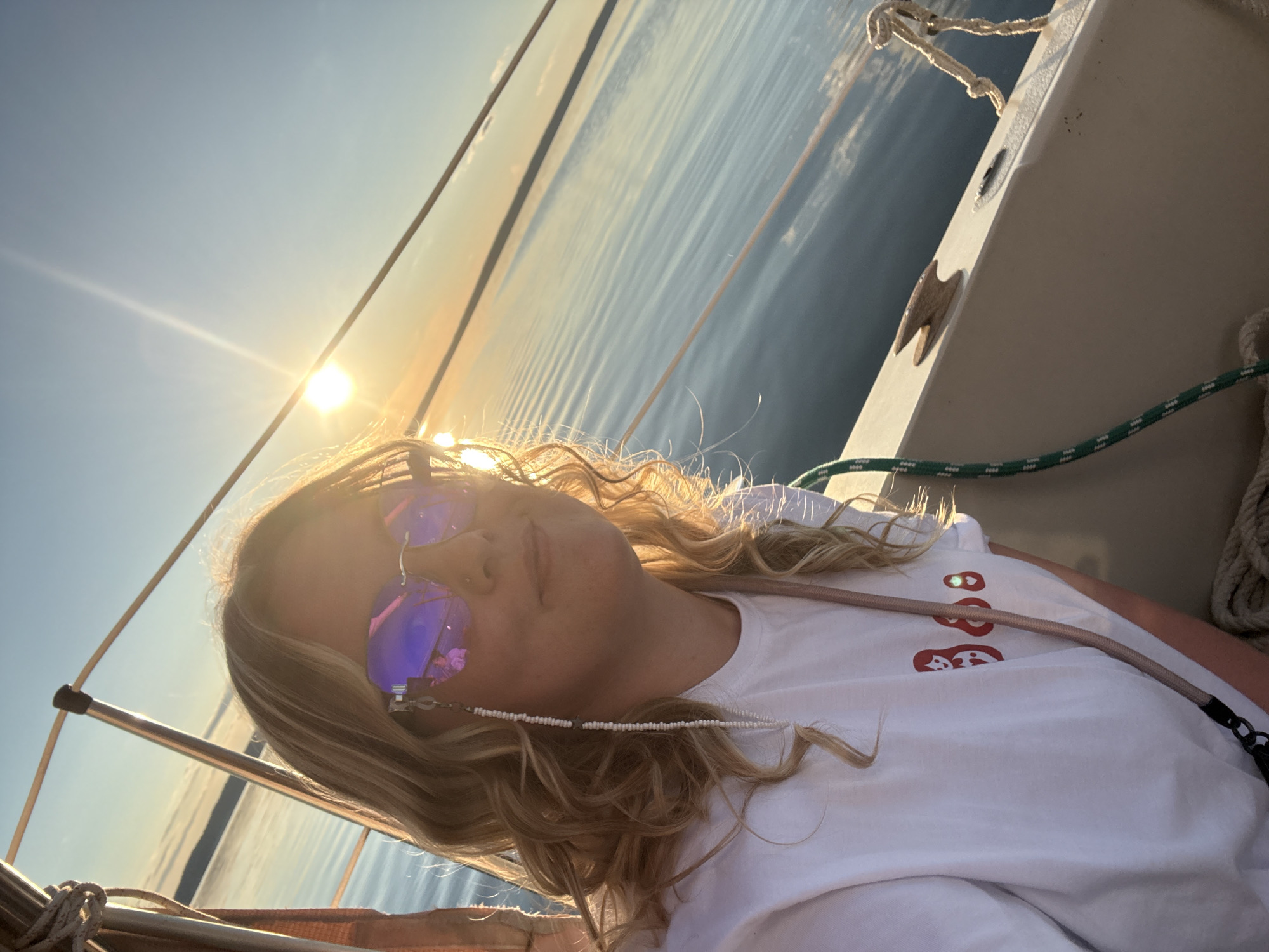 An overcast selfie taken from a low angle shows a young, blonde woman with wavy hair and sunglasses on a sailboat. She is wearing a white t-shirt with red matryoshka doll graphics and a beaded chain attached to her sunglasses. The sun is setting over a body of water, casting a warm glow on the scene. The water is calm with small ripples. The boat's railing and rigging are visible in the foreground. The image is taken in Chaykovsky, Russia.