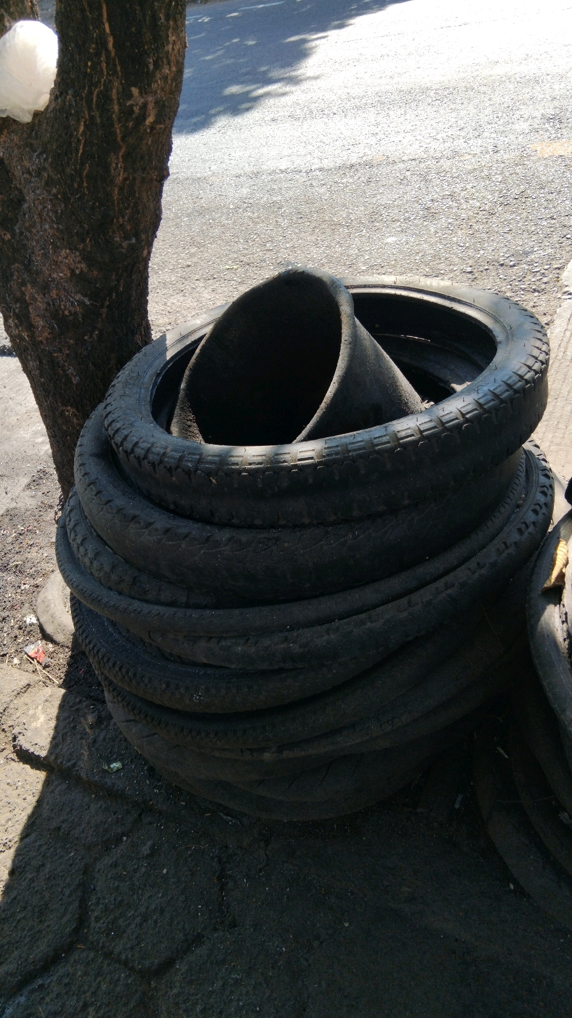 A stack of black tires, some interlocked and one partially collapsed inwards, rests on a dirt and asphalt surface next to the base of a tree. The tree trunk, on the left, is rough-barked and dark. A white object, possibly plastic sheeting, is draped over the upper part of the tree trunk. The background shows a grey asphalt road with some dappled sunlight and shadow from unseen foliage. The scene appears to be outdoors, possibly a roadside or a service area in Proyonanggan Tengah, Indonesia.