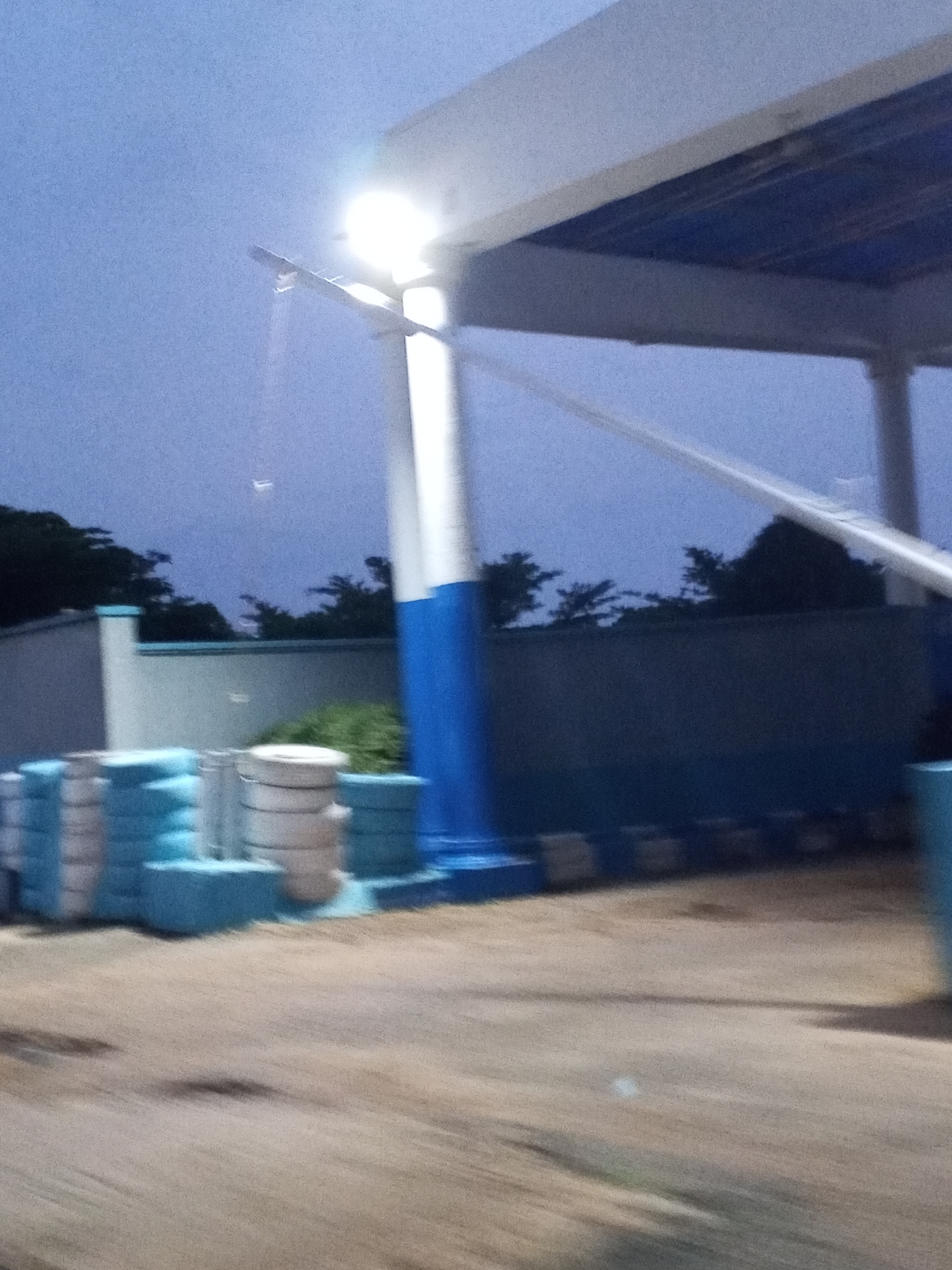 A blurred, low-light, eye-level exterior shot taken in Jos, Nigeria, depicts a covered structure with a white roof and blue underside, supported by a tall, white and blue striped pillar. A bright light emanates from a fixture above. In the foreground, stacks of what appear to be light blue and white cylindrical objects, possibly tires or traffic barriers, are arranged. A grey wall, possibly part of a building or enclosure, runs horizontally across the middle ground. Silhouetted trees and foliage are visible in the background, against a twilight sky. The overall impression is one of a dimly lit, perhaps partially constructed or storage area, under an overhanging structure, with a hazy, indistinct foreground and background due to motion blur and low light conditions.