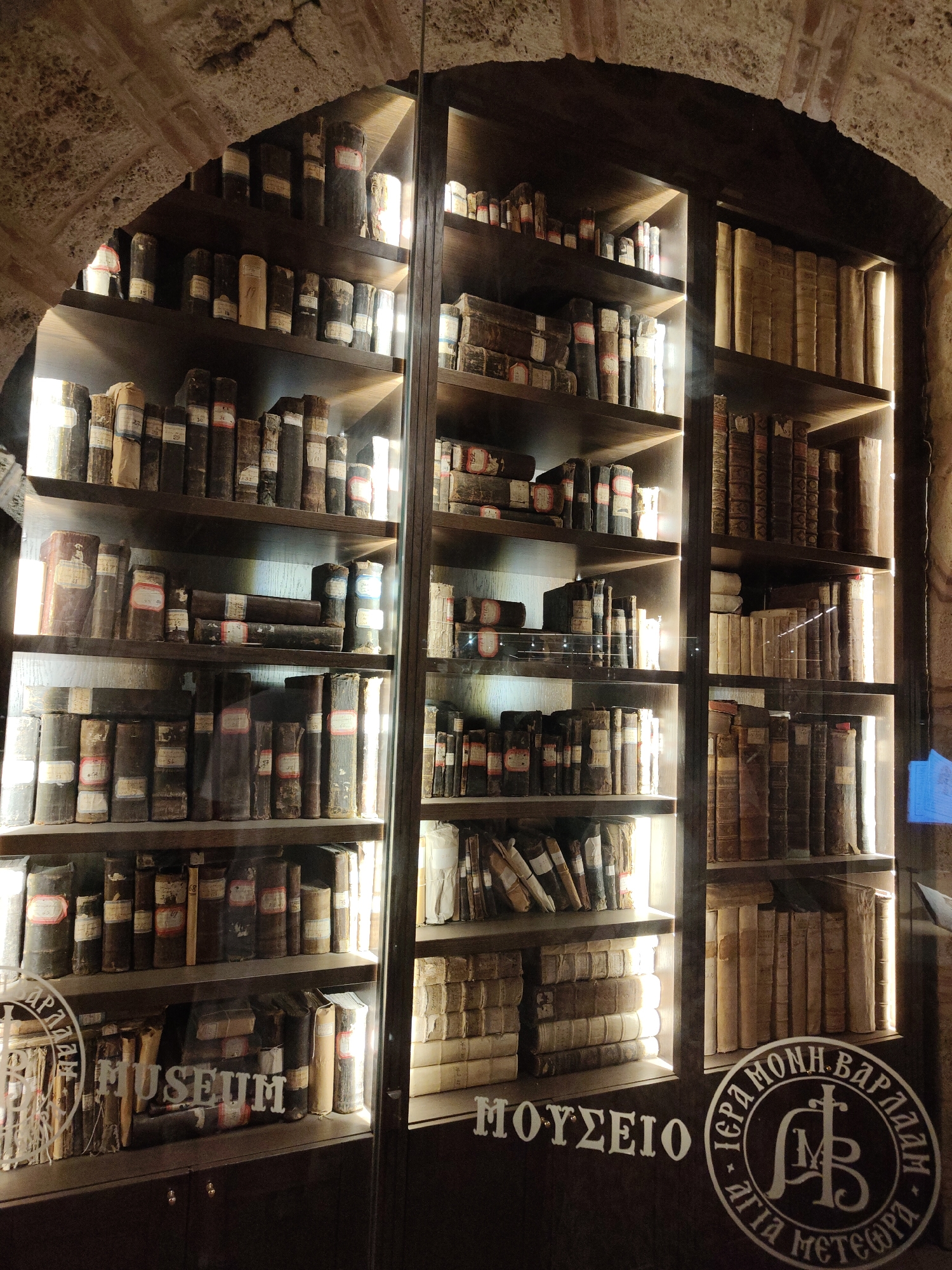 The image shows an interior view of a glass-enclosed bookshelf filled with numerous old books, located within the Holy Monastery of Great Meteoron in Kalambaka, Greece. The monastery is a UNESCO World Heritage site renowned for its historical and religious significance, situated atop towering rock formations. The bookshelf is made of dark wood with multiple shelves, illuminated from behind, creating a warm glow that highlights the spines of the ancient volumes. Many of the books appear to be leather-bound, with some having visible labels or markings. The environment suggests a museum or archival setting within the monastery, preserving these historical texts. Visible text in Greek on the glass includes "ΜΟΥΣΕΙΟ" (Museum) and a circular emblem with text relating to "ΙΕΡΑ ΜΟΝΗ ΒΑΡΛΑΑΜ" (Holy Monastery of Varlaam), indicating the location's connection to the Meteora monastic community. The architectural elements of the monastery, such as the arched stone ceiling visible in the upper left, are also present. There are no people or actions depicted in the image; it is a still scene focused on the display of books.