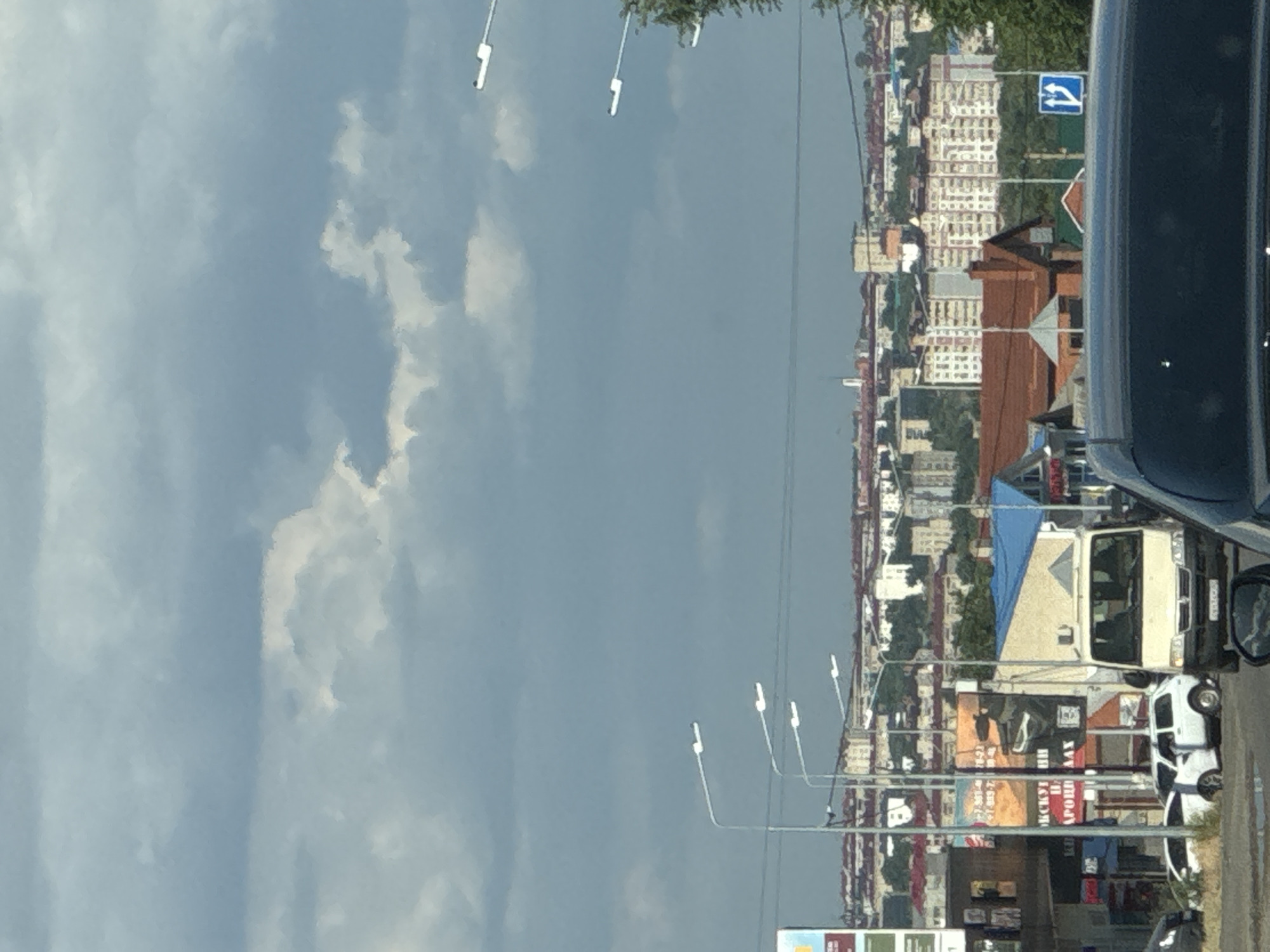 This is a sideways, eye-level shot of a street scene in Dorozhny, Russia, likely taken from inside a vehicle. The image is dominated by a cloudy sky on the left and a city street with buildings and vehicles on the right. The time of day appears to be daytime, given the brightness, though the heavy cloud cover suggests it might be overcast or approaching rain.
On the right side of the frame, a variety of buildings can be seen, ranging from multi-story apartment complexes with cream-colored facades to smaller, residential structures with red-tiled roofs. There is a prominent white panel van parked on the street, partially covered by a blue tarp. Several cars are also visible, including a white hatchback. A traffic sign indicating a mandatory turn is present. Streetlights on poles line the road.
There are several billboards and signs with Russian text visible along the street. One billboard has large, colorful graphics and what appears to be a phone number. Another sign includes the Cyrillic words "РОССИЙСКИЙ" and "НАРОДНЫЙ", suggesting a public or national theme.
The overall impression is that of a typical urban environment on a cloudy day. There are no people clearly visible in the frame, but the presence of vehicles and buildings indicates human activity and habitation. The composition is dynamic due to the sideways perspective, making it feel like a candid snapshot of everyday life.