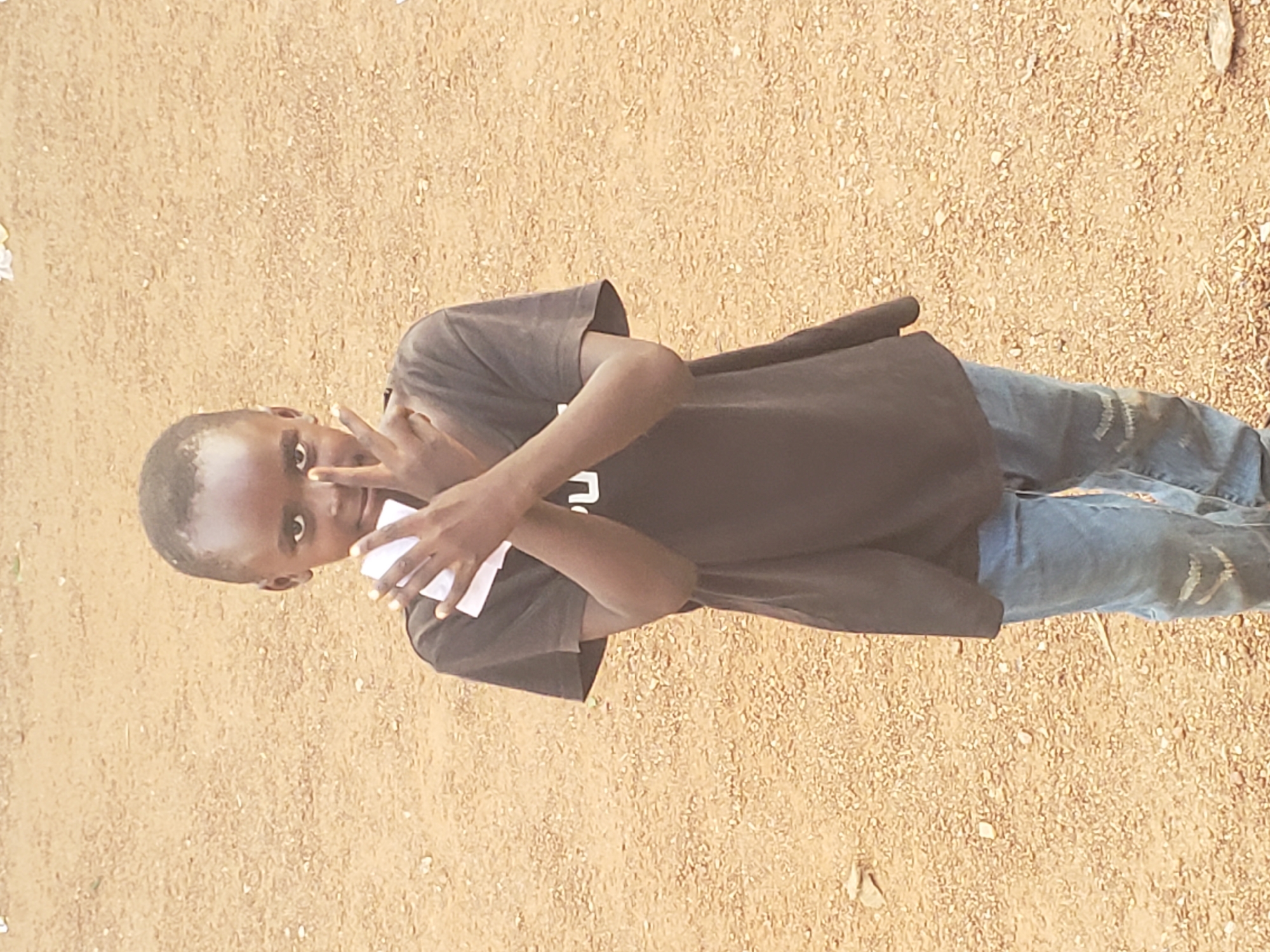 A young boy stands centrally in an outdoor setting in Gumel, Nigeria, looking directly at the viewer. He has dark skin and short dark hair. He wears a dark brown or black short-sleeved t-shirt with a partially visible white letter "U" or "PU" on the chest, and light blue denim jeans that appear faded and slightly distressed, particularly around the knees. His hands are raised and crossed in front of his face. His right hand is positioned over his left, with the index and middle fingers extended into a "V" shape, touching his face. Both hands hold small, white, rectangular objects, resembling pieces of paper or thin cards. The ground beneath him and extending into the background is an unpaved, reddish-brown sandy or gravelly surface, scattered with small stones and light-colored debris. The scene is illuminated by natural daylight, without harsh shadows.