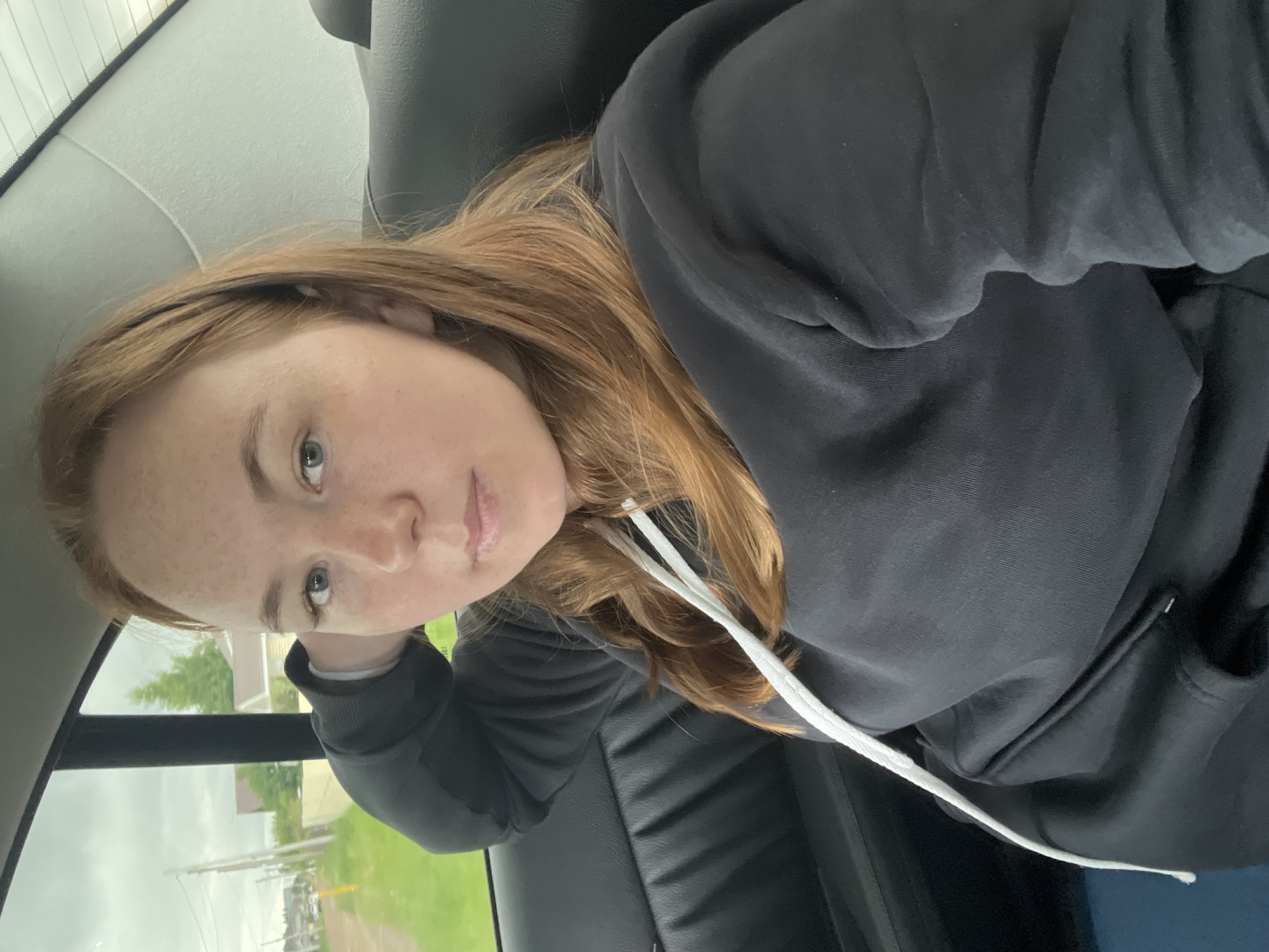 The image features a young adult female occupying the rear passenger seat of a vehicle. She has long, reddish-brown hair, fair skin with prominent freckles across her nose and forehead, and blue-grey eyes. She is wearing a dark grey or black hooded sweatshirt with white drawstrings and maintains a neutral expression while looking directly at the camera. Her left arm is bent, with her hand resting behind her head against the dark-colored interior panel of the car. The vehicle's upholstery appears dark, likely leather or vinyl, and the roof lining is light-colored with faint horizontal lines visible at the top right.
Through the car's side window on the left, an exterior scene is visible: an unpaved road, green grass and trees, multiple utility poles, and distant residential structures under an overcast sky. The image itself provides no visual identifiers to confirm the specific location as Прикамский, Russia.