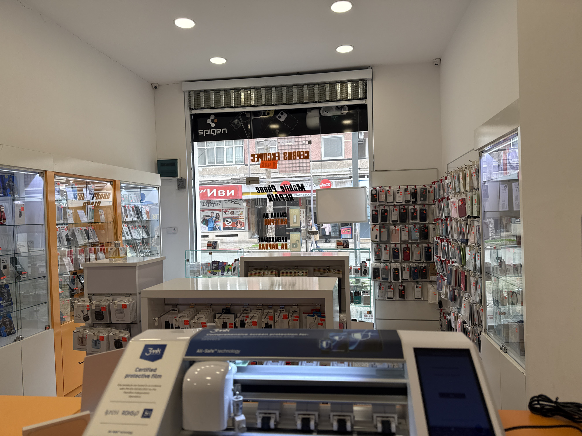 Here's a description of the scene:
The image captures the interior of a mobile phone accessory store. The location is inferred to be in Burgas, Bulgaria, based on the Cyrillic script visible on storefront signs through the shop window.
The store is well-lit with overhead fluorescent lights. A large display window shows the street outside, with various businesses and buildings visible. Brands like "Spigen" are displayed on the inside of the window. Shelves and glass display cases are stocked with phone cases, screen protectors, and other accessories. A protective film plotter device branded "3mk All-Safe technology" is situated prominently in the foreground, presumably for cutting custom screen protectors. Display stands and tables are organized neatly throughout the store. A security camera is mounted near the top of the frame.