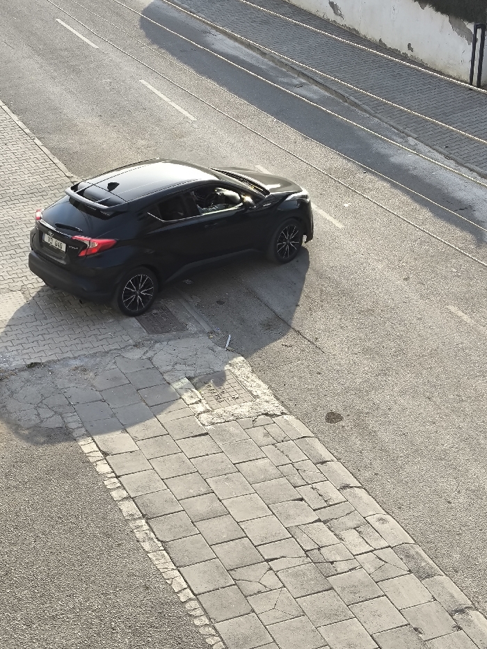 The image provides an elevated view of a street scene in Nicosia, Northern Cyprus / Cyprus. A black Toyota C-HR, displaying the Northern Cyprus license plate "YD 040," is positioned diagonally across a curb. Its front-left and rear-left wheels are on the asphalt road, while its front-right and rear-right wheels are on a grey paved sidewalk. A human figure is visible in the driver's seat, facing left.
The asphalt road features faded white double solid lines and a single white dashed line further right. A small, dark, circular stain is present on the asphalt near the sidewalk. The sidewalk is constructed from rectangular grey paving blocks, exhibiting numerous cracks, uneven surfaces, and areas of spalling. A metal utility cover, partially obscured, is embedded in the sidewalk, displaying faint text that includes "TEL-KO." White debris, possibly paper, is scattered on the sidewalk near the utility cover. Beyond the road, a weathered, light-colored wall with two dark vertical posts is visible. Strong sunlight casts distinct shadows of the vehicle and other elements towards the lower-right, indicating clear daytime conditions.