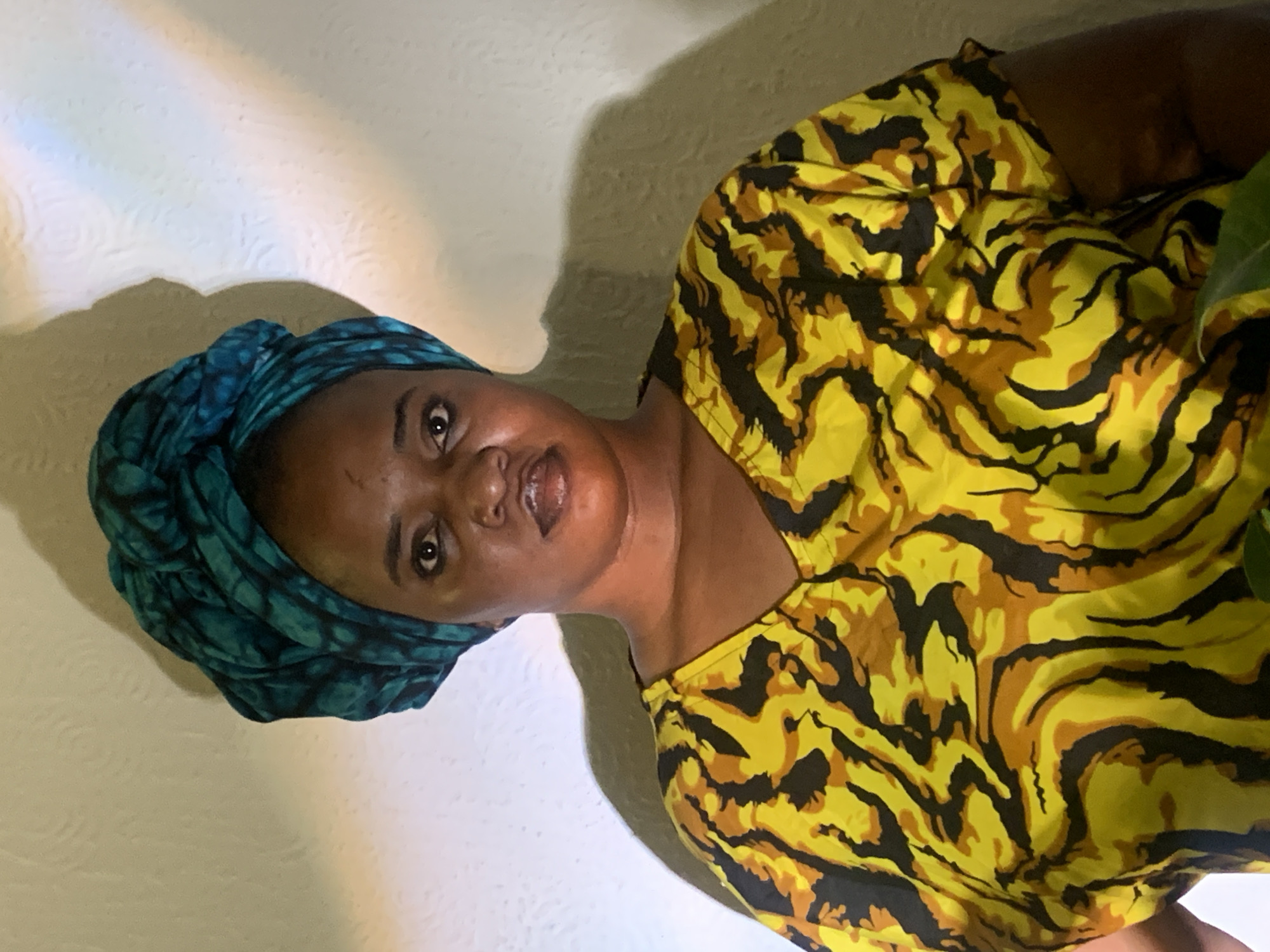 An indoor portrait captured in Barakin Akawo, Nigeria, features an individual with dark skin, positioned against a light-colored, textured wall. The person wears a vibrant, intricately patterned teal and black headwrap, paired with a yellow, brown, and black abstract-patterned top that evokes an animal print. The individual gazes directly at the viewer with a neutral to subtly pleasant expression. Shadows are visible on the wall behind them, suggesting specific lighting conditions, possibly artificial, or natural light filtering in. A very faint, colorful light refraction, resembling a small rainbow, is subtly present on the upper left wall. A hint of green foliage from a plant is partially visible on the right side of the frame. The subject's head is tilted slightly to the left within the composition. No text is visible in the image.