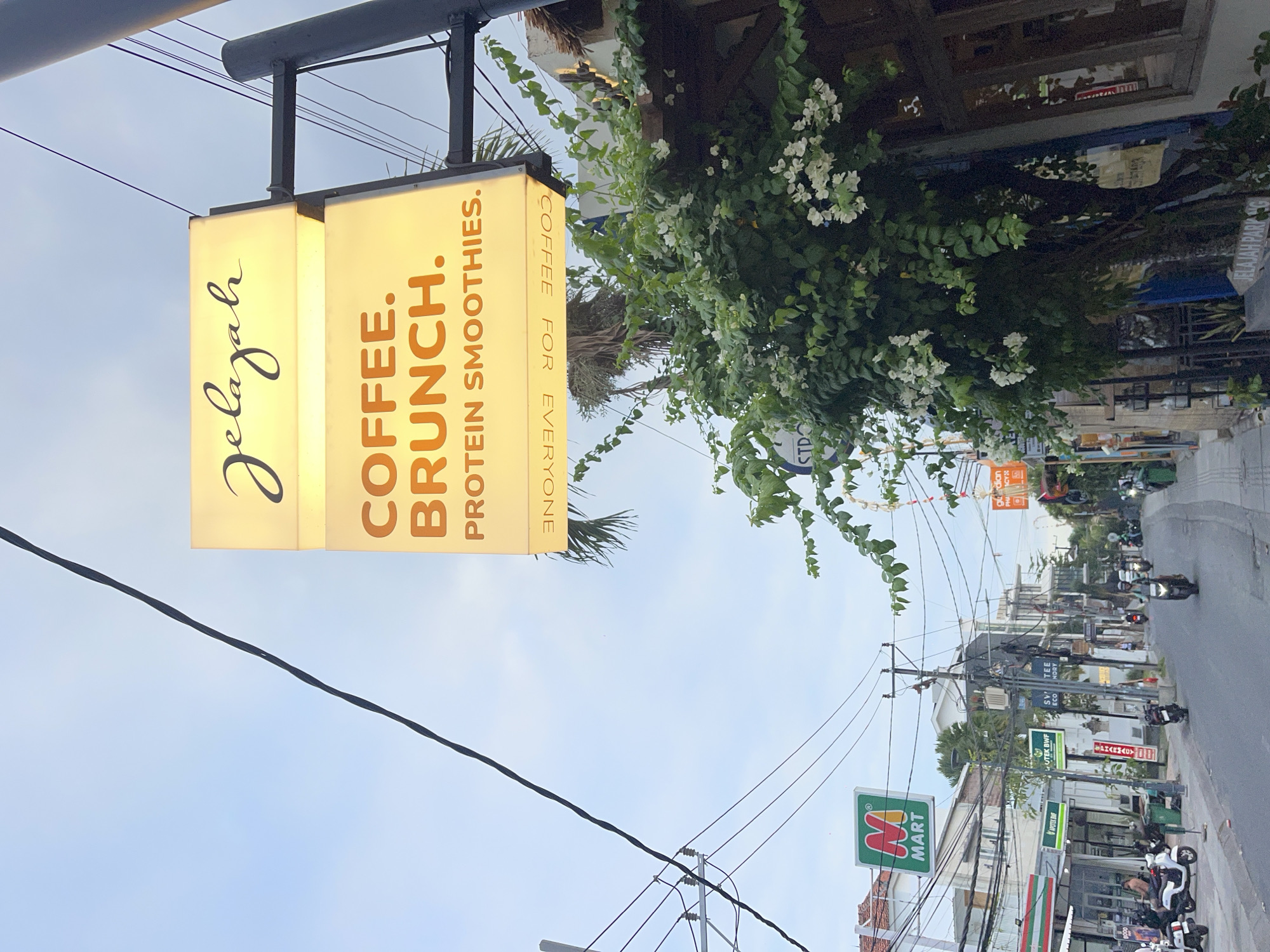 The image captures a street scene in North Kuta, Indonesia, with a focus on a brightly lit sign for "Jelajah" which advertises "Coffee, Brunch, Protein Smoothies". The sign is mounted on a pole, with a lower, smaller sign facing the street displaying "COFFEE FOR EVERYONE" in reverse. The background shows a street lined with various establishments, including what appears to be a minimart labeled "M MART" with green and red signage. Several motorbikes are parked along the sidewalk and a few are on the street, with some riders visible. Lush green foliage with white flowers spills over a wall in the foreground, partially obscuring a building with wooden-framed windows. Utility poles with numerous wires crisscross the sky, typical of the urban landscape. The sky is overcast, suggesting it is either early morning or late afternoon.