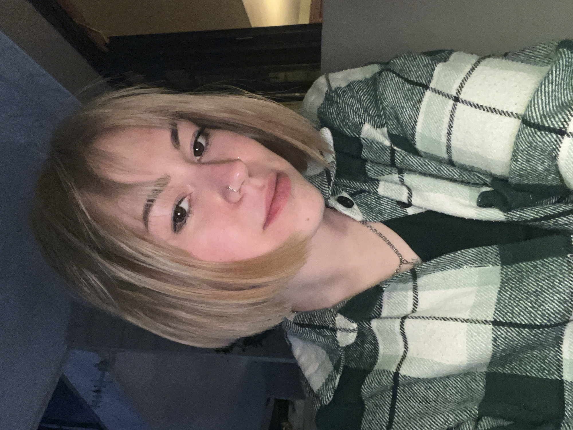 A medium indoor shot depicts a young female with short, blonde hair and brown eyes. She has a nose ring in her left nostril. She is wearing a green and white plaid flannel shirt over a dark top. A thin silver necklace is visible. The background shows a partially visible window or picture frame. The location is not discernible from the image.