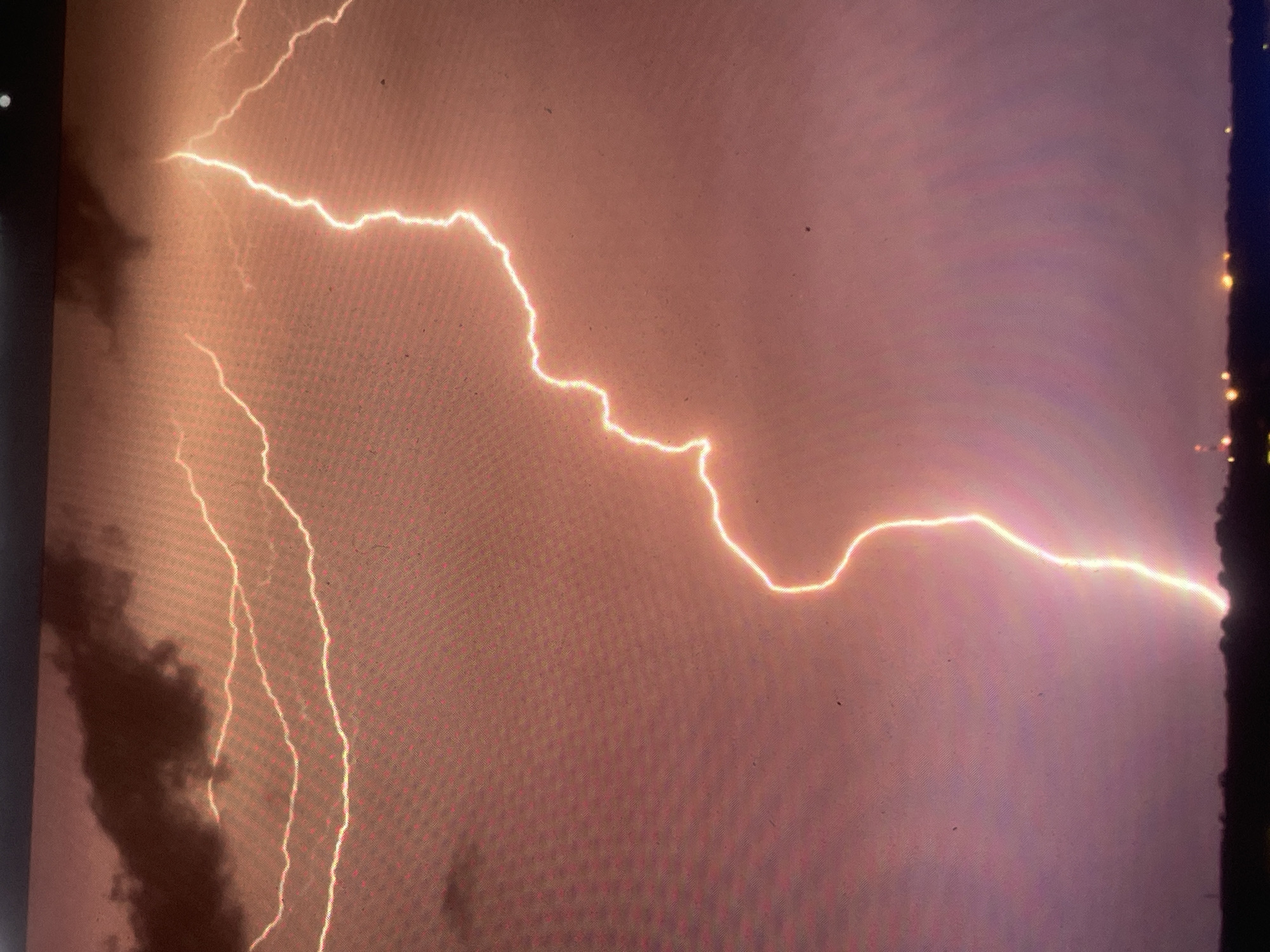 The image displays a prominent lightning strike illuminating a stormy night sky over Zelenyi Hai, Ukraine. A singular, intensely bright, and highly jagged lightning bolt originates from the upper-right cloud mass and descends diagonally, connecting with the distant ground line in the lower-center. Multiple thinner, horizontal lightning flashes are embedded within the dark, dense cloud cover occupying the upper-left quadrant. The sky is characterized by deep reddish-orange to purple coloration, indicative of the atmospheric conditions during the electrical discharge. A dark, silhouetted landmass, appearing as a distant treeline or low hills, forms the horizon across the lower portion of the frame. Scattered small, yellow-orange artificial lights are visible below this horizon, suggesting distant infrastructure or a settlement. No individuals or specific objects are identifiable on the landmass. The overall image exhibits a pixel grid pattern, consistent with a photograph taken of a digital screen, and includes minor dust particles or screen imperfections.