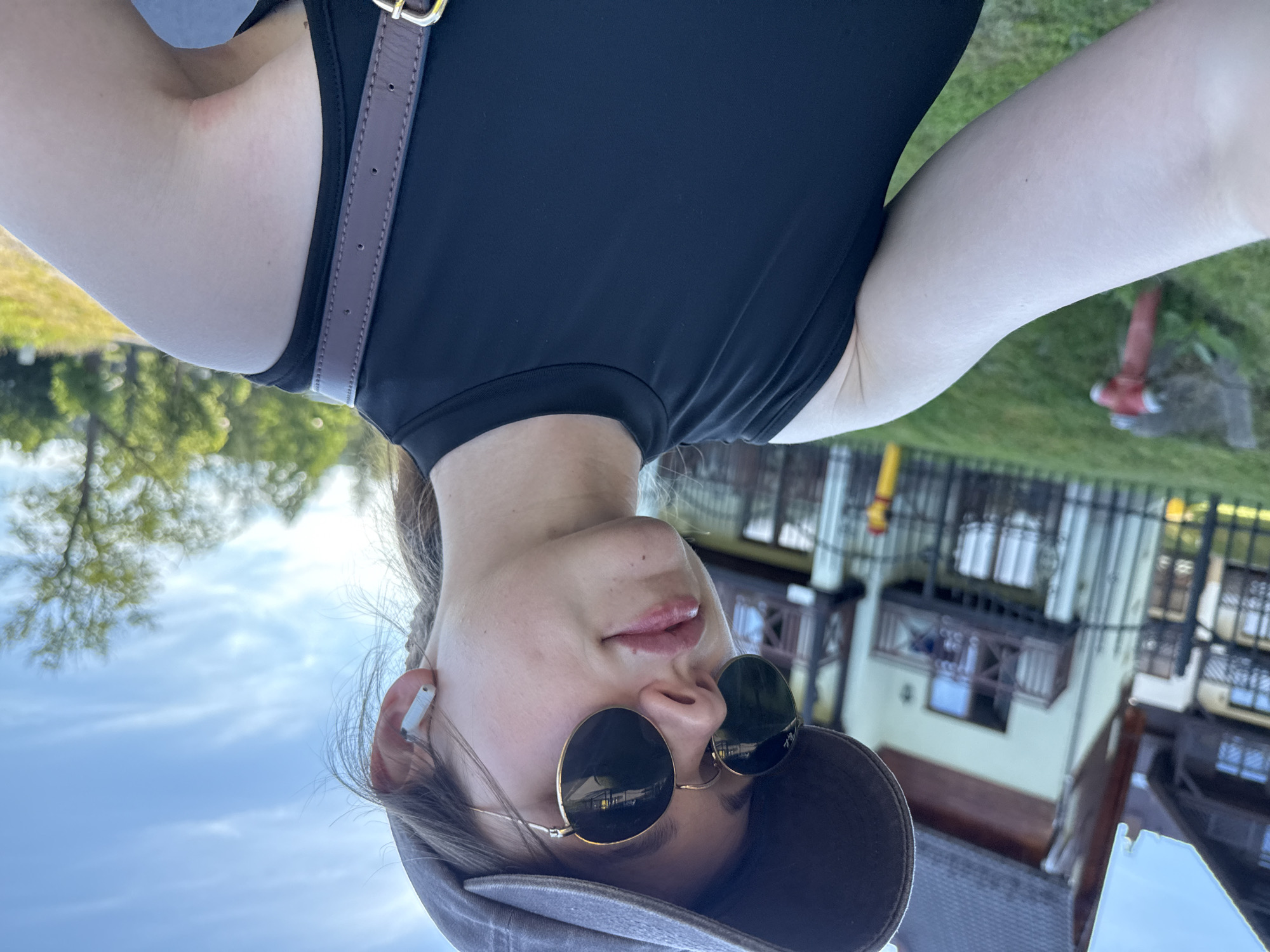 A young Caucasian woman wearing a grey baseball cap, round sunglasses, and a black sleeveless top is outdoors, in what appears to be a residential area with a modern apartment building in the background. She is also wearing white wireless earbuds. The building has wooden balconies and is surrounded by green grass. A red fire hydrant is visible on the left side of the image. The sky is blue with some wispy clouds. The lighting suggests it is daytime, possibly late afternoon given the angle of the sun. The image was taken in Szklarska Poręba, Poland.