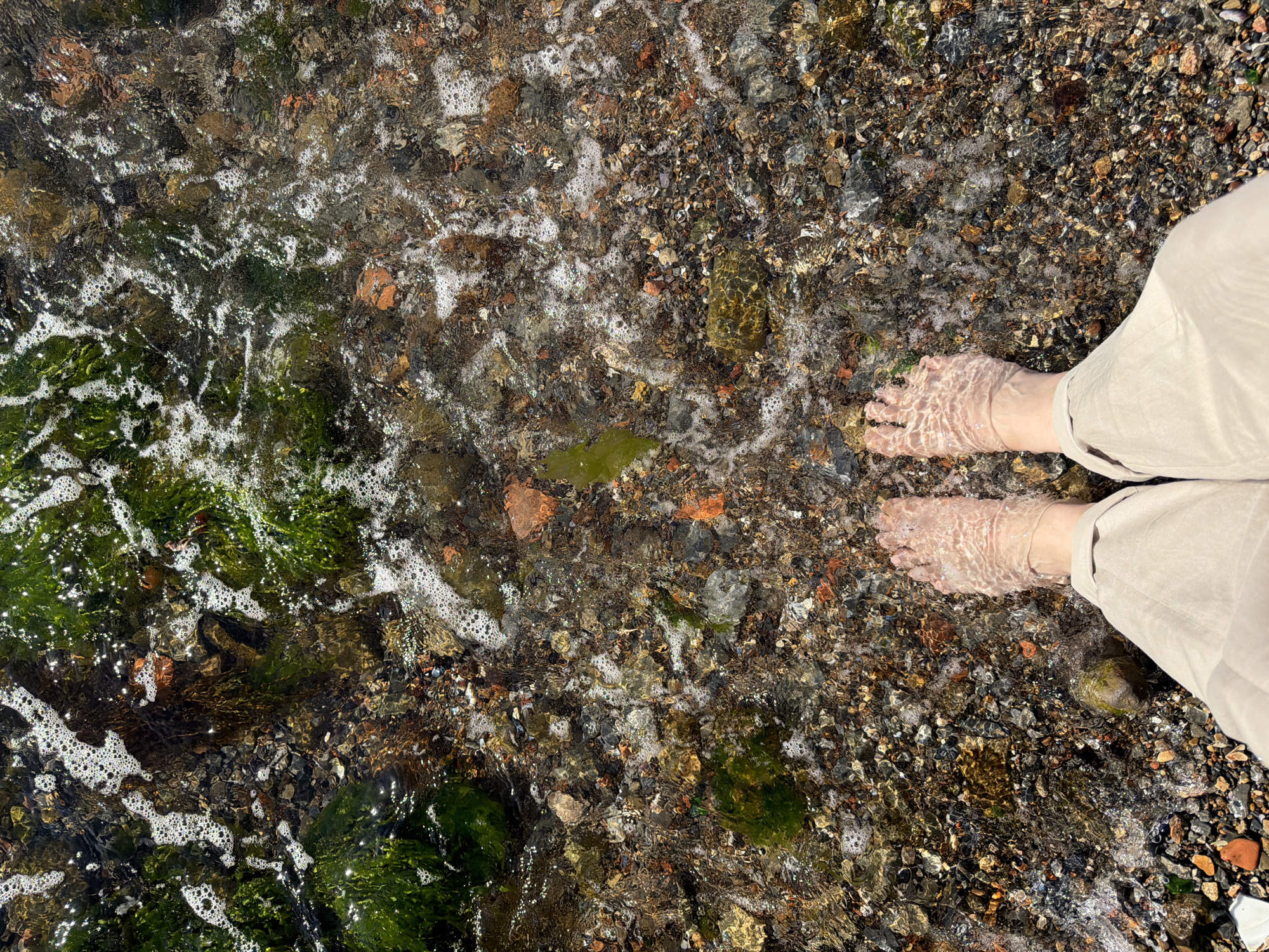 The image shows a top-down view of a person's feet submerged in shallow, clear water on a pebble beach. The water is gently lapping, creating small waves and foam around the feet. The pebbles on the beach are varied in color and size, with some small pieces of green seaweed visible. The person is wearing light-colored pants that extend down to their ankles. The lighting suggests it is daytime. The specific location of Üsküdar, Turkey, cannot be determined from the visual information provided in the image.