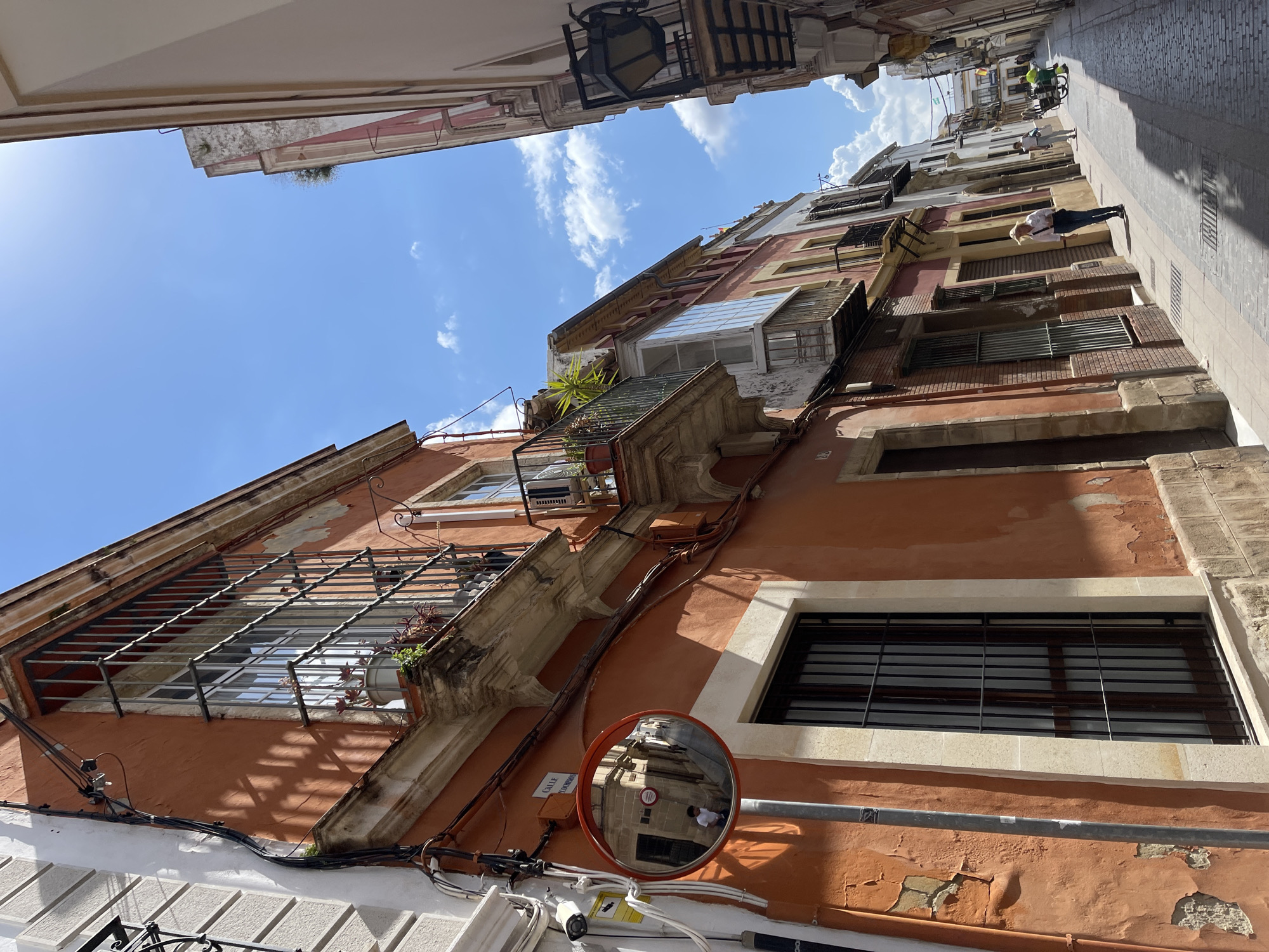 The image depicts a narrow street scene in Jerez De La Frontera, Spain. The perspective is looking up at the buildings, revealing their aged facades painted in shades of orange and beige. Architectural details include window bars, balconies, and protruding ledges. Electrical wiring runs along the building's exterior. A convex safety mirror is affixed to the wall, reflecting a section of the street and a pedestrian. Above, the sky is visible with light clouds. Other pedestrians can be seen walking down the street. The buildings stand close together, creating a confined urban space.