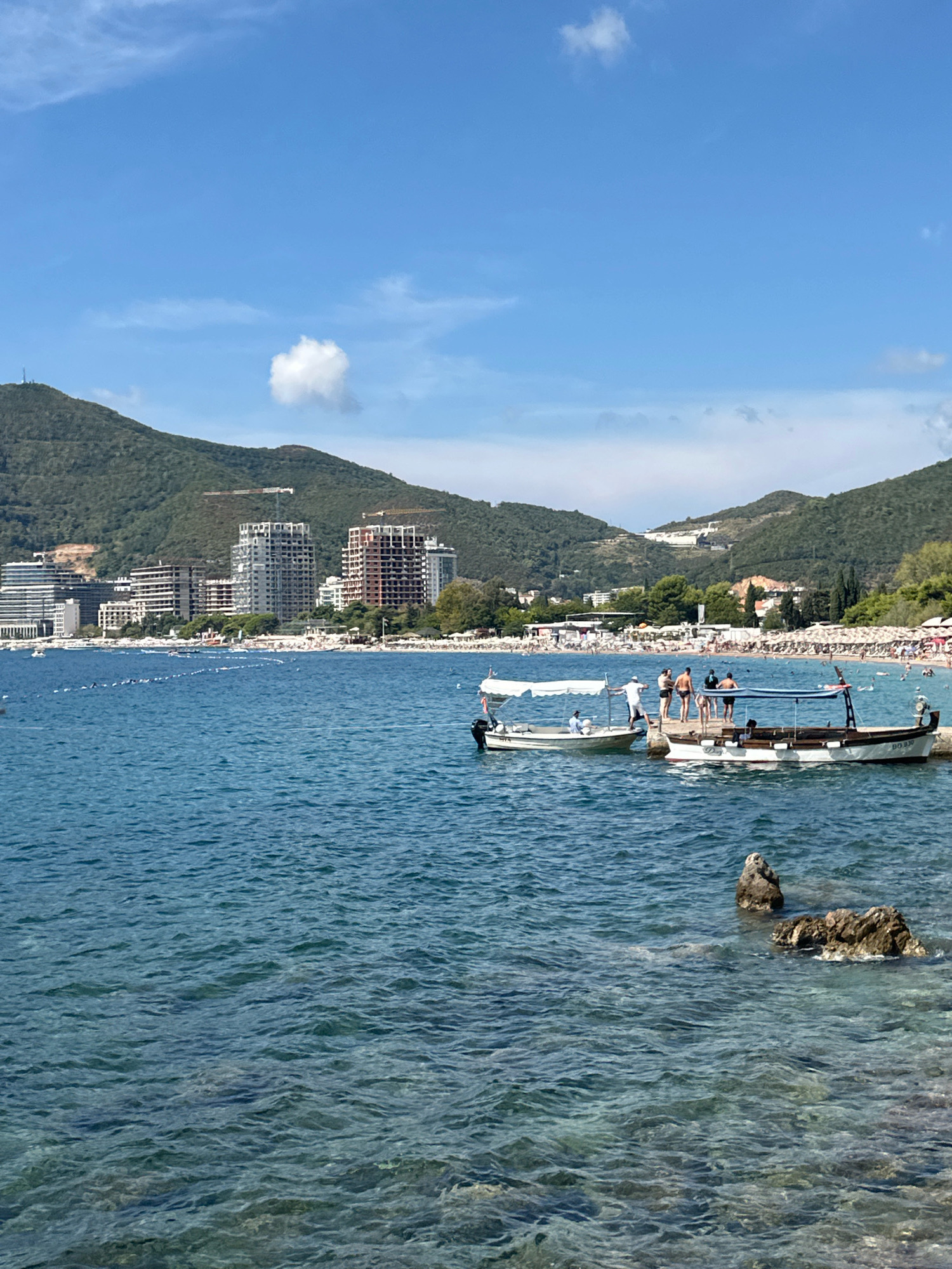In Budva, Montenegro, on a clear, sunny day, the turquoise waters of the Adriatic Sea gently lap against a rocky shore. In the foreground, two small boats are docked at a pier. The boat on the left is white with a canvas awning and an outboard motor. The boat on the right is larger, also white with wooden accents, and features a distinctive dark-colored stern with a registration number. Several individuals are visible on the pier, some interacting with each other and the boats, including one person appearing to be a boat operator.
Beyond the pier, a bustling beach is visible, lined with numerous umbrellas and sunbathers. The coastline stretches along the bay, backed by verdant, hilly terrain. Several modern buildings, some appearing to be under construction with visible cranes, are nestled into the hillsides. The sky above is a vibrant blue with a few scattered white clouds, contributing to the serene and picturesque coastal scene.