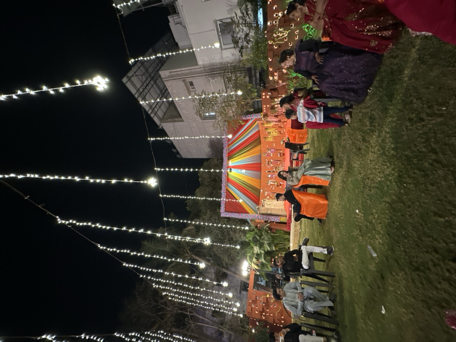 Outdoor nighttime event featuring string lights. A vibrant backdrop, presumably for a ceremony or stage, is the focal point. People are gathered on the lawn, some seated and others standing. Several individuals are dressed in formal attire or traditional garments. The event seems festive, set against a backdrop of a building.