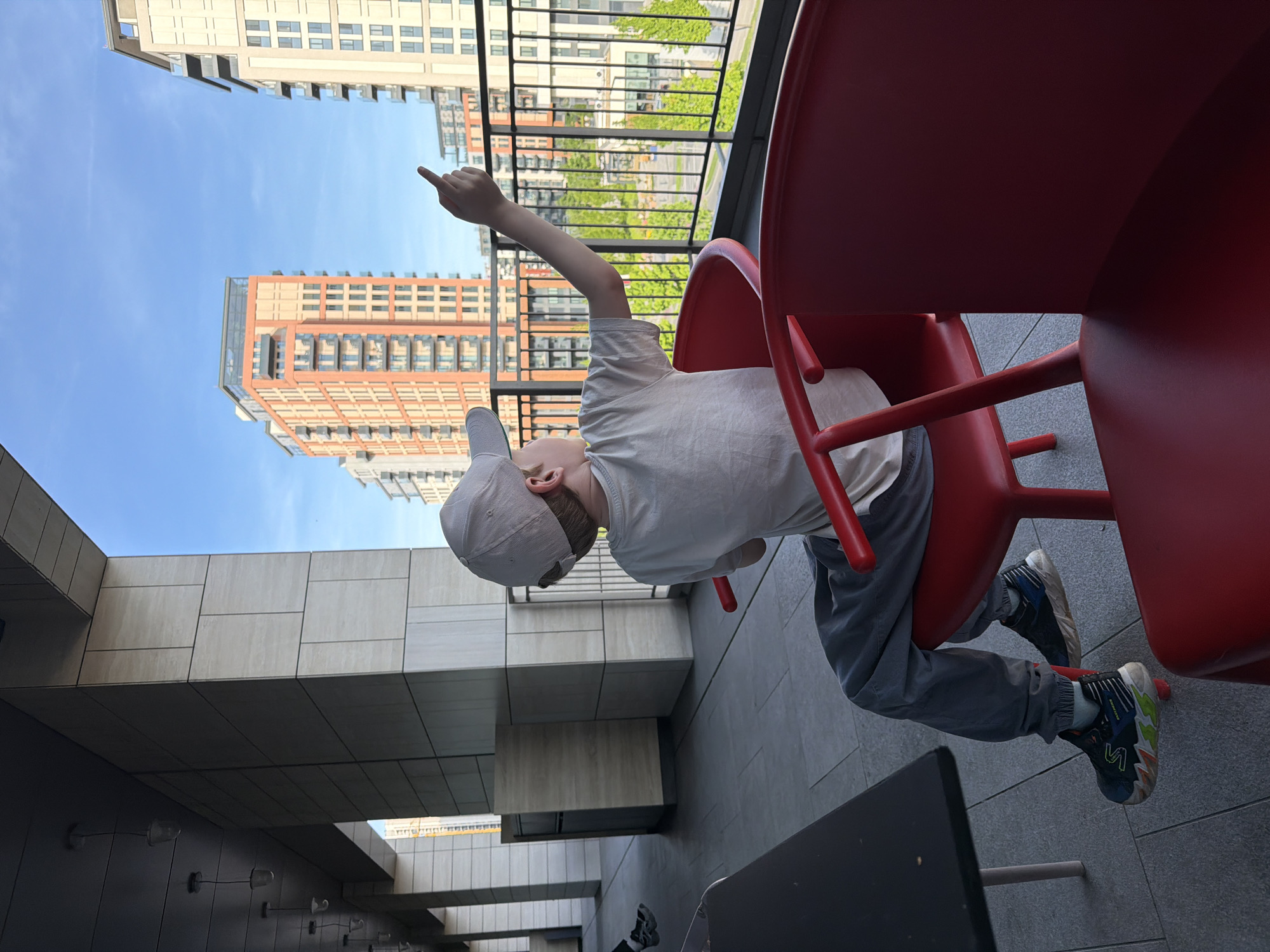 A boy in a white t-shirt, gray sweatpants, a gray baseball cap, and athletic shoes sits in a red chair on a gray-tiled balcony. He points his right arm upwards towards a tall building with beige and red brick on a blue sky. Behind him, metal railings overlook greenery. The building with the boy is on the left, showing light-colored rectangular panels. A black table is in the foreground on the lower right. The location is in Belgrade, Serbia.